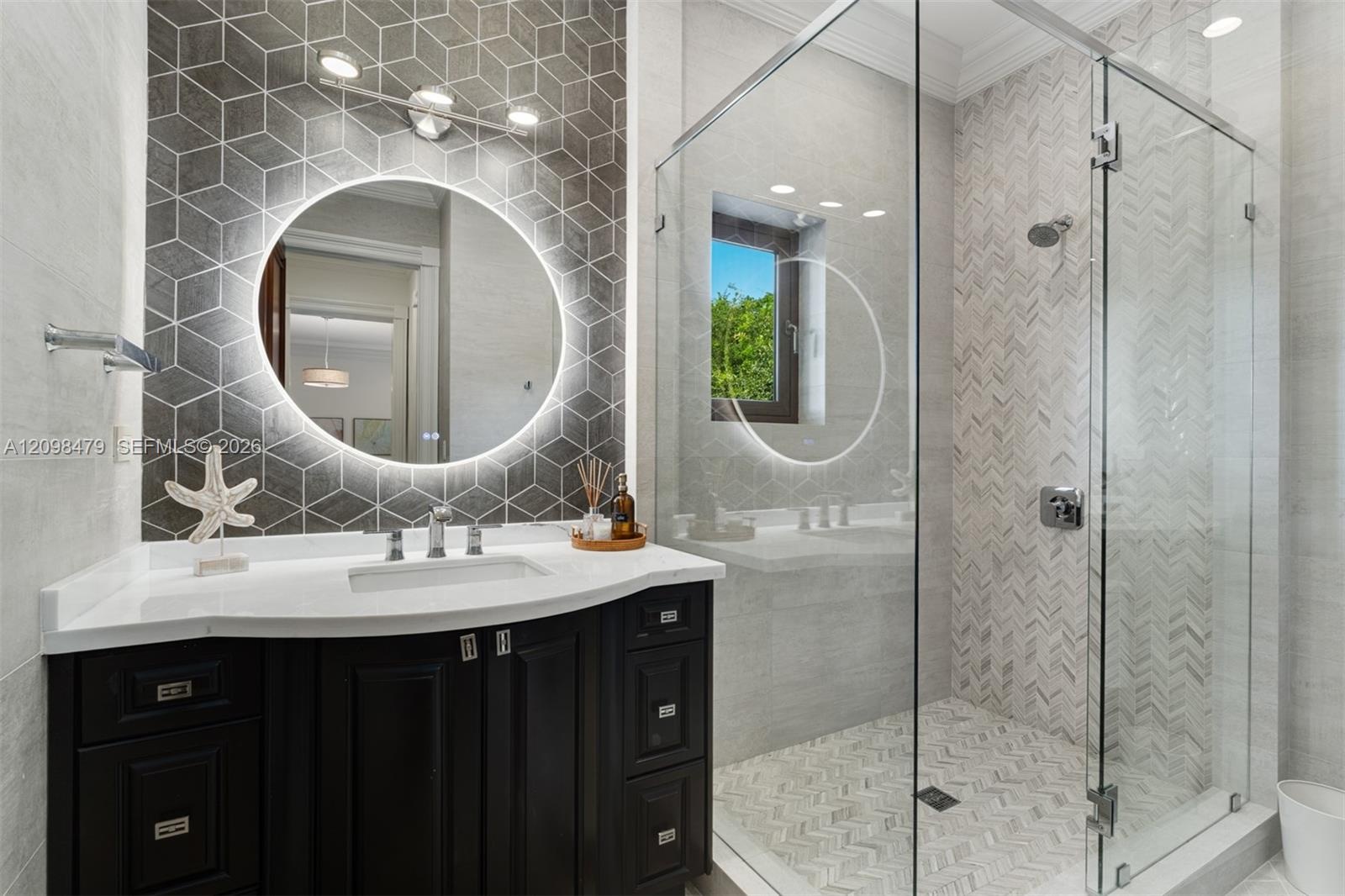 Vanity with integrated sink and chrome fixtures, illuminated round mirror, geometric wall tile, glass-enclosed shower, and herringbone tile shower sur