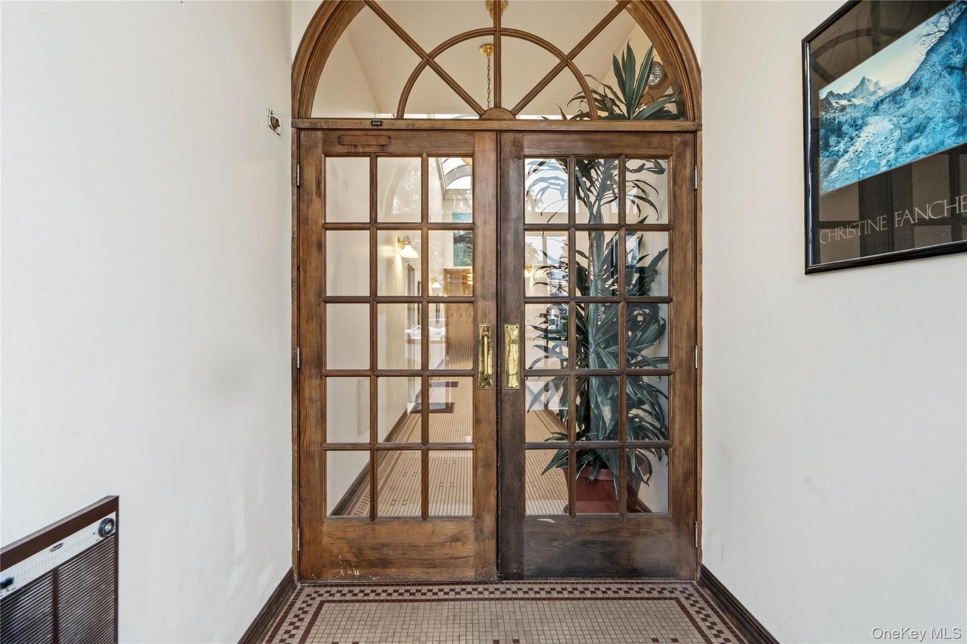 Double wood-framed glass entry doors featuring a decorative arched transom, multi-pane glazing, and brass hardware, accented by tiled flooring with a 