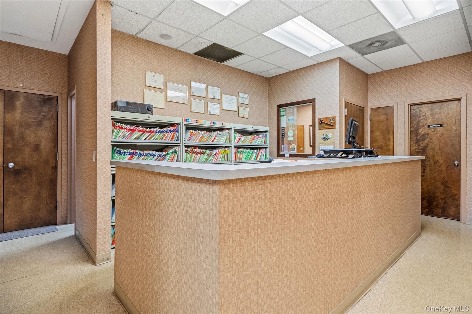 Reception area featuring an L-shaped service counter, suspended ceiling tiles with integrated lighting, two solid wood doors, and built-in shelving un