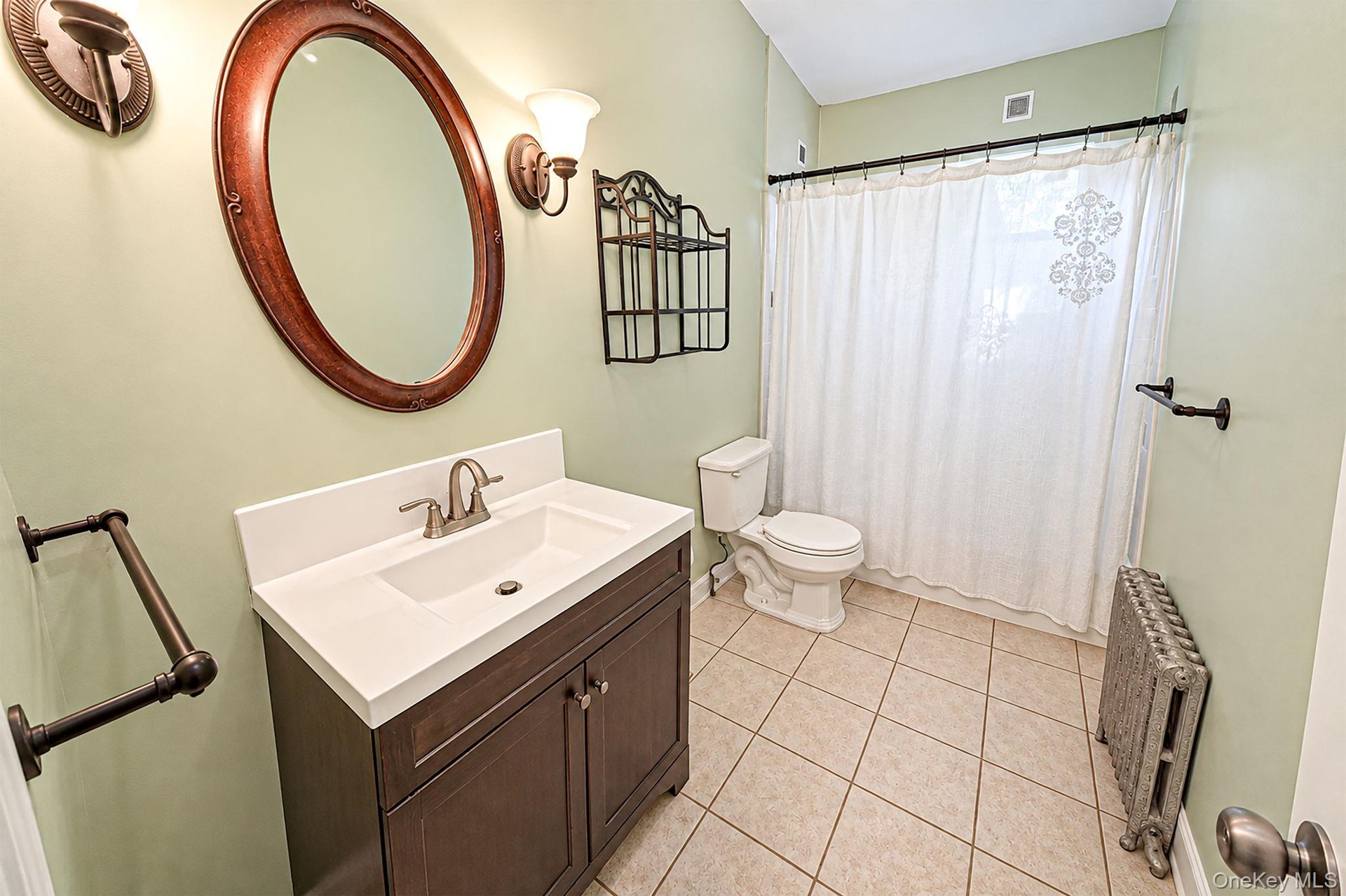 Dark-finish wooden vanity with a white countertop and integrated sink, neutral tile flooring, oval wall mirror, and dual wall-mounted light fixtures
