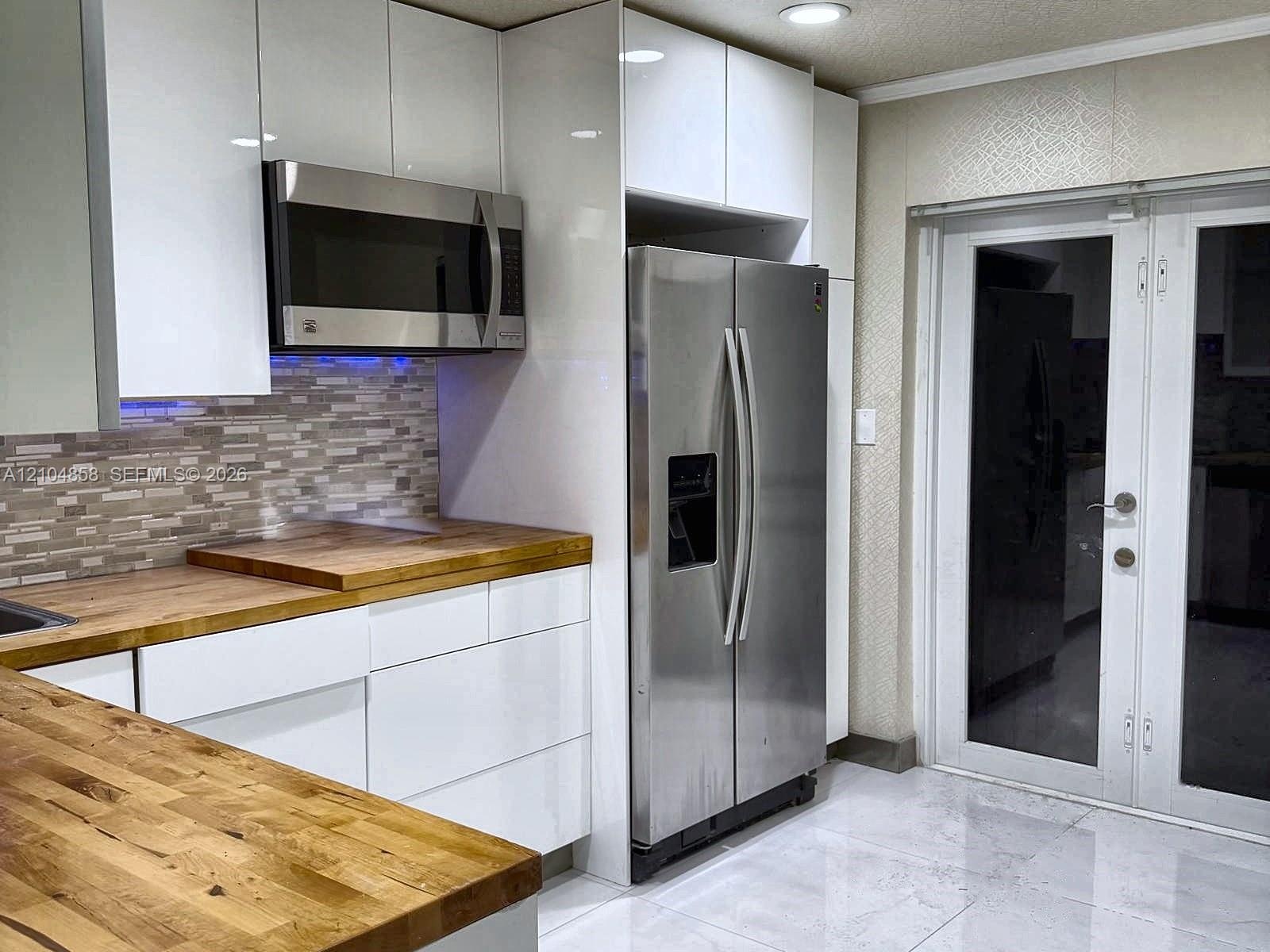 Kitchen featuring high-gloss white cabinetry, wood-finish countertops, stainless steel appliances, multi-tonal tile backsplash, and French doors