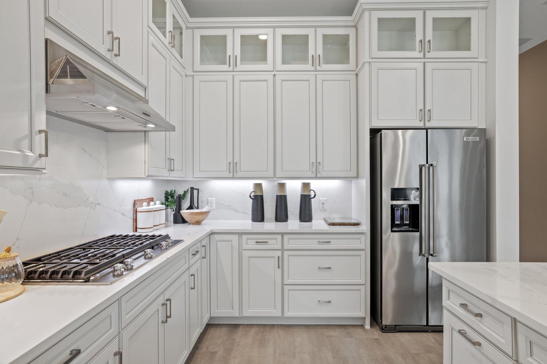 White shaker-style cabinetry with glass-front uppers, light-toned stone-finish countertops, full-height stone-finish backsplash, stainless steel gas c