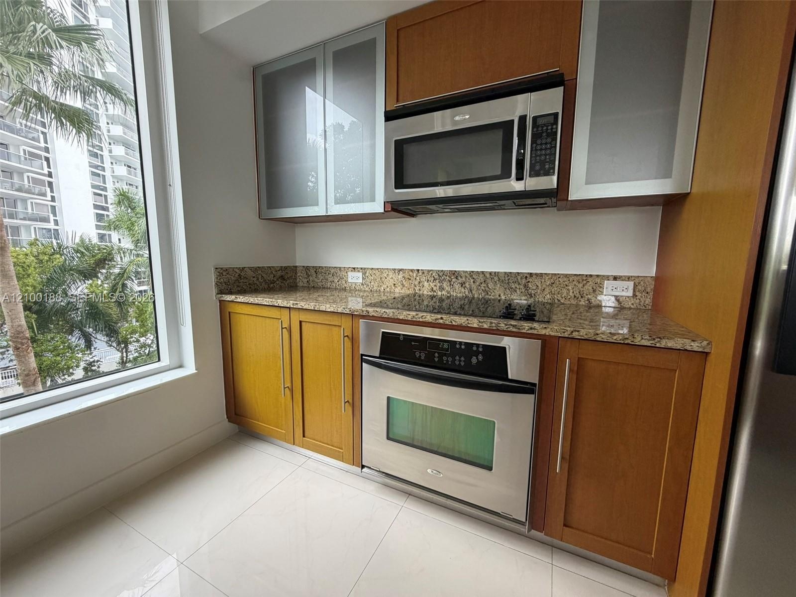 Kitchen featuring wood cabinetry, stone countertops, stainless steel built-in appliances, glass-panel upper cabinet doors, and large-format tile floor