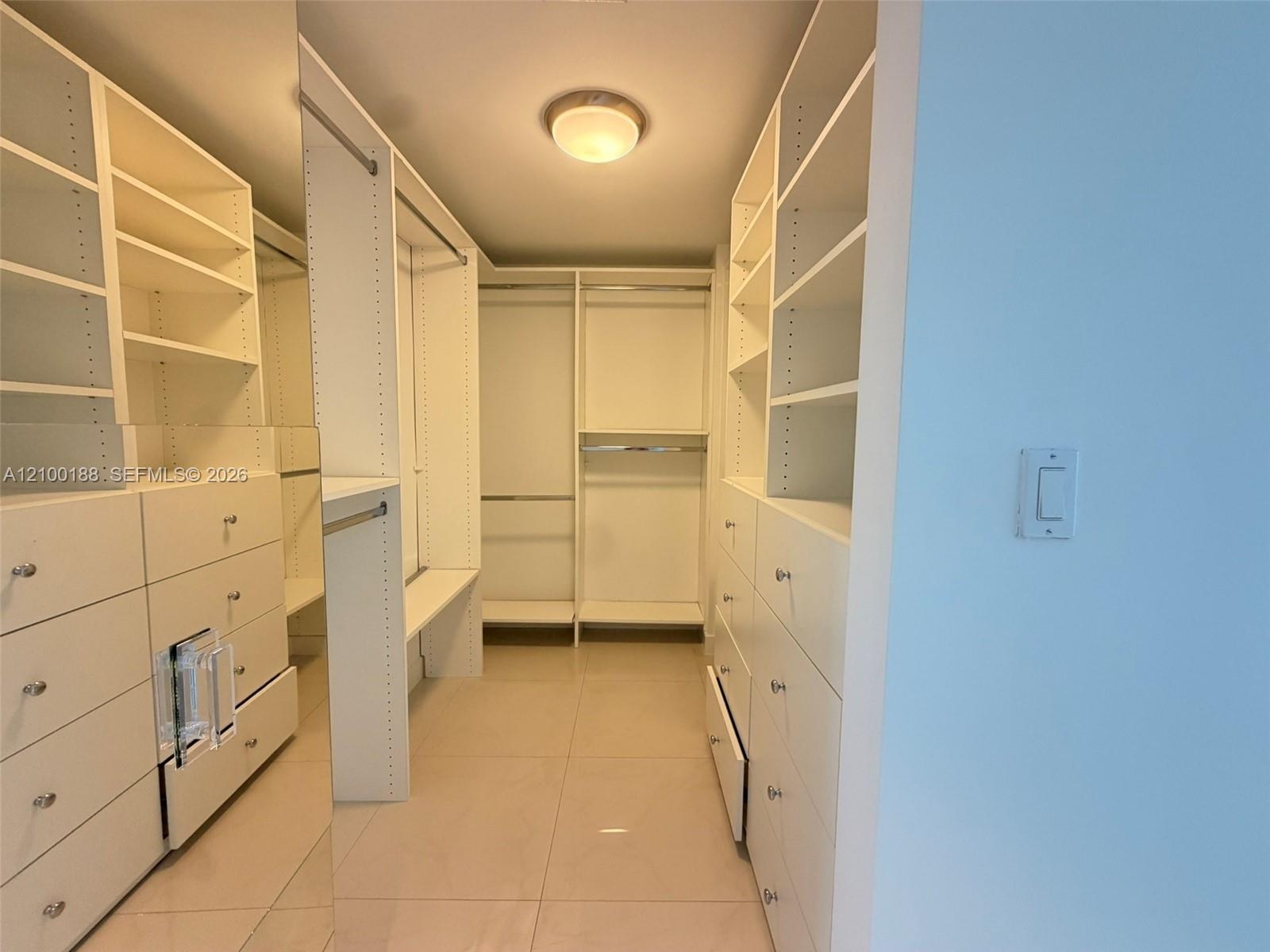 Custom walk-in closet featuring white built-in shelving, floor-to-ceiling cabinetry, integrated hanging rods, and neutral tile flooring
