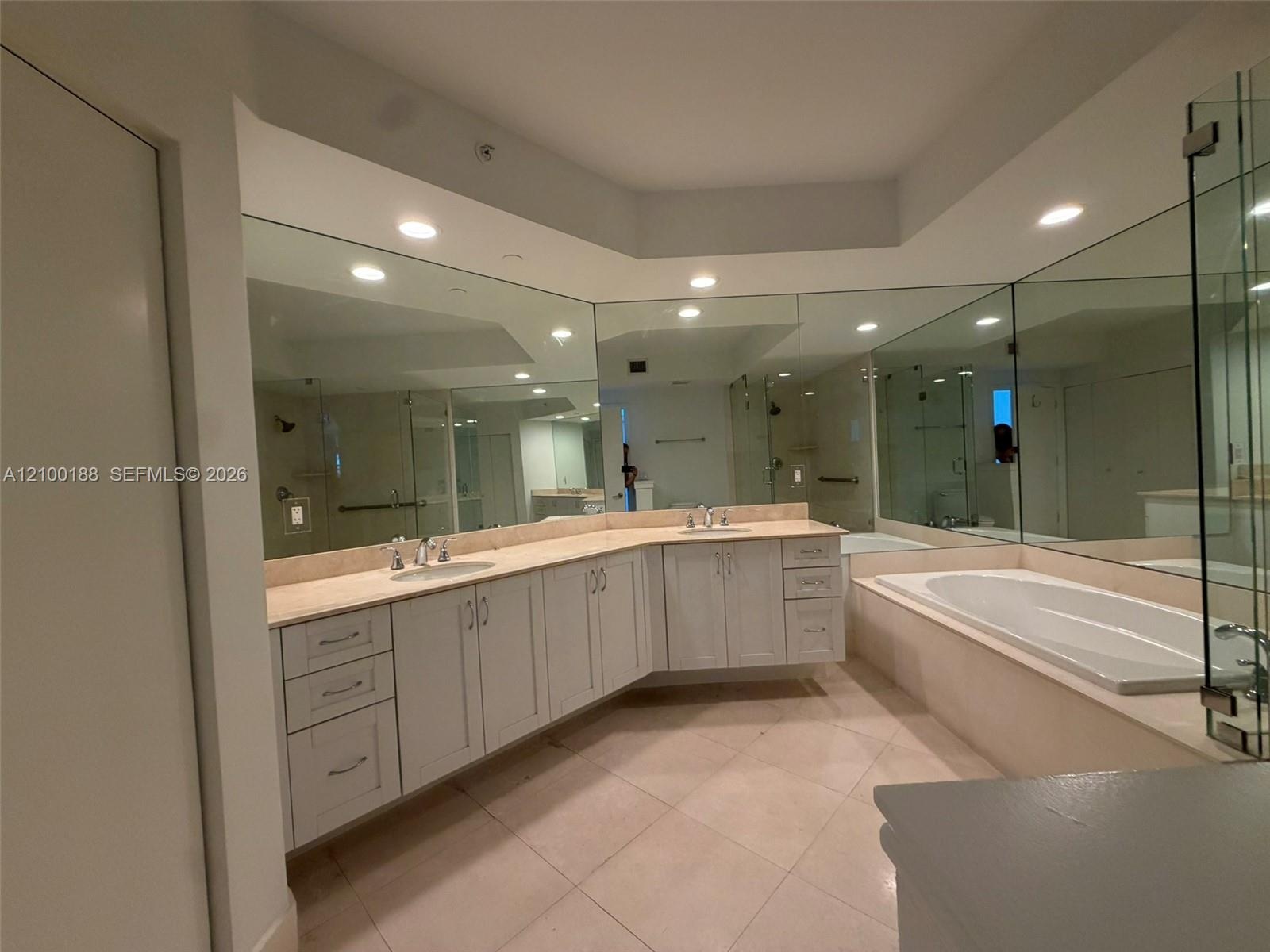 Dual vanity featuring light-toned cabinetry, a neutral countertop, recessed lighting, expansive wall mirrors, and an integrated soaking tub