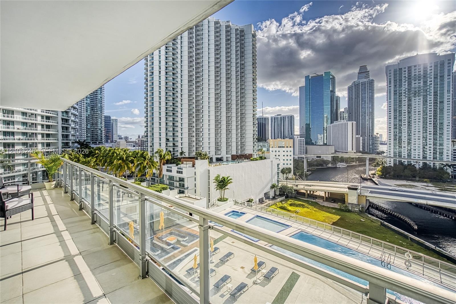 Covered balcony featuring tile flooring and a glass-paneled railing overlooking an urban skyline and multi-level swimming pools