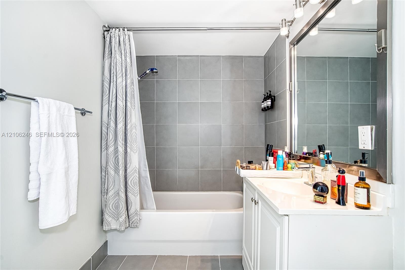 Bathroom featuring a white vanity with a rectangular sink, wall-to-wall mirror, chrome light fixture, and a shower-tub combination with large-format g