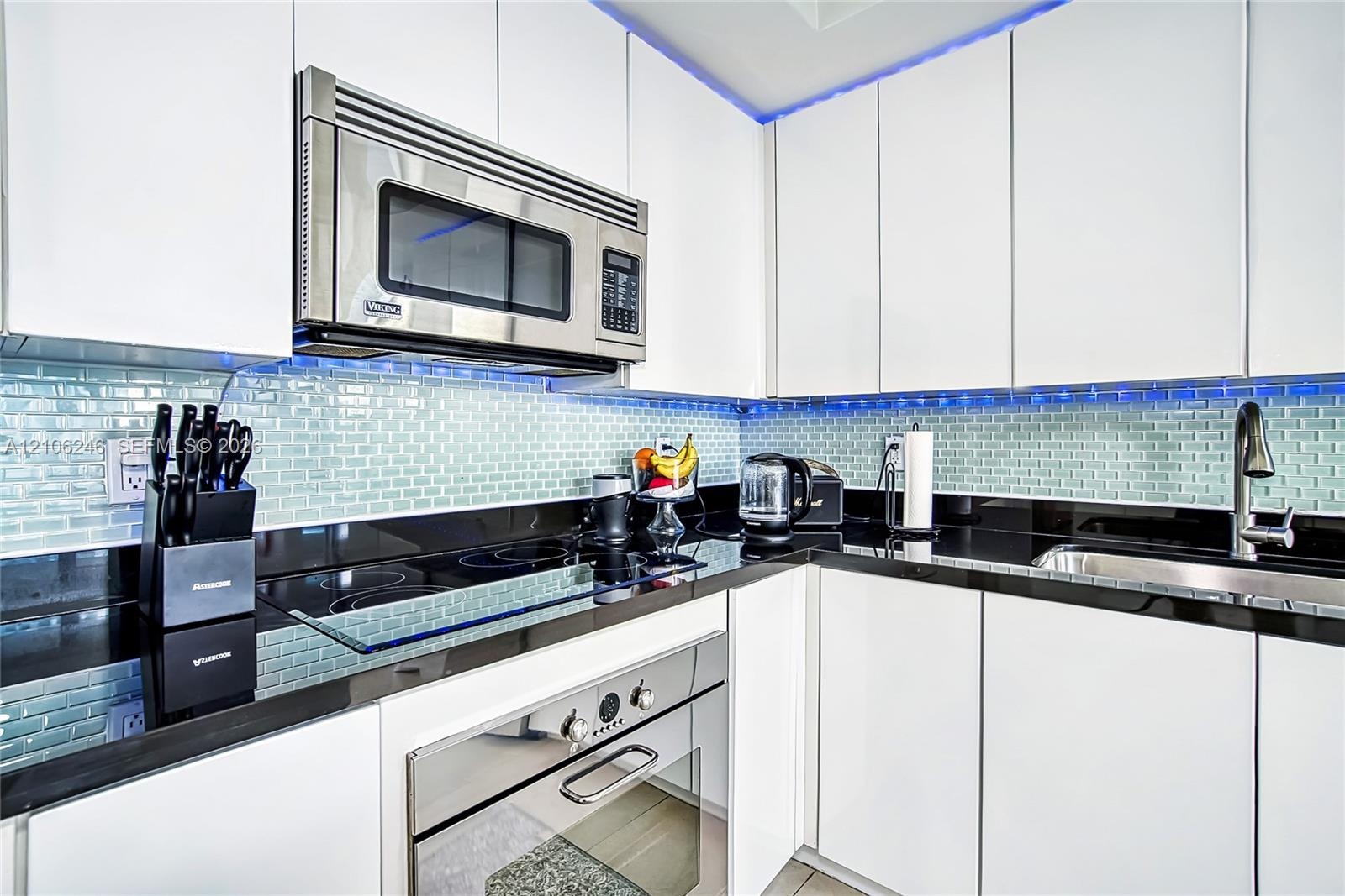 Modern kitchen featuring flat-panel white cabinetry, contrasting black countertops, glass subway tile backsplash, stainless steel built-in microwave, 