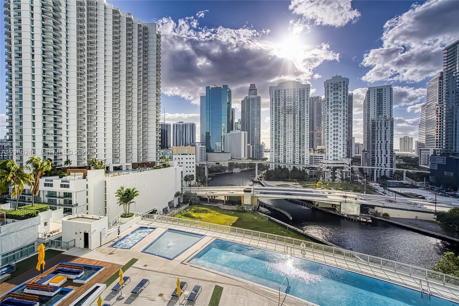 Outdoor pool terrace featuring tiered swimming areas, sun deck with stone-finish tiling, glass panel perimeter railing, and urban skyline backdrop