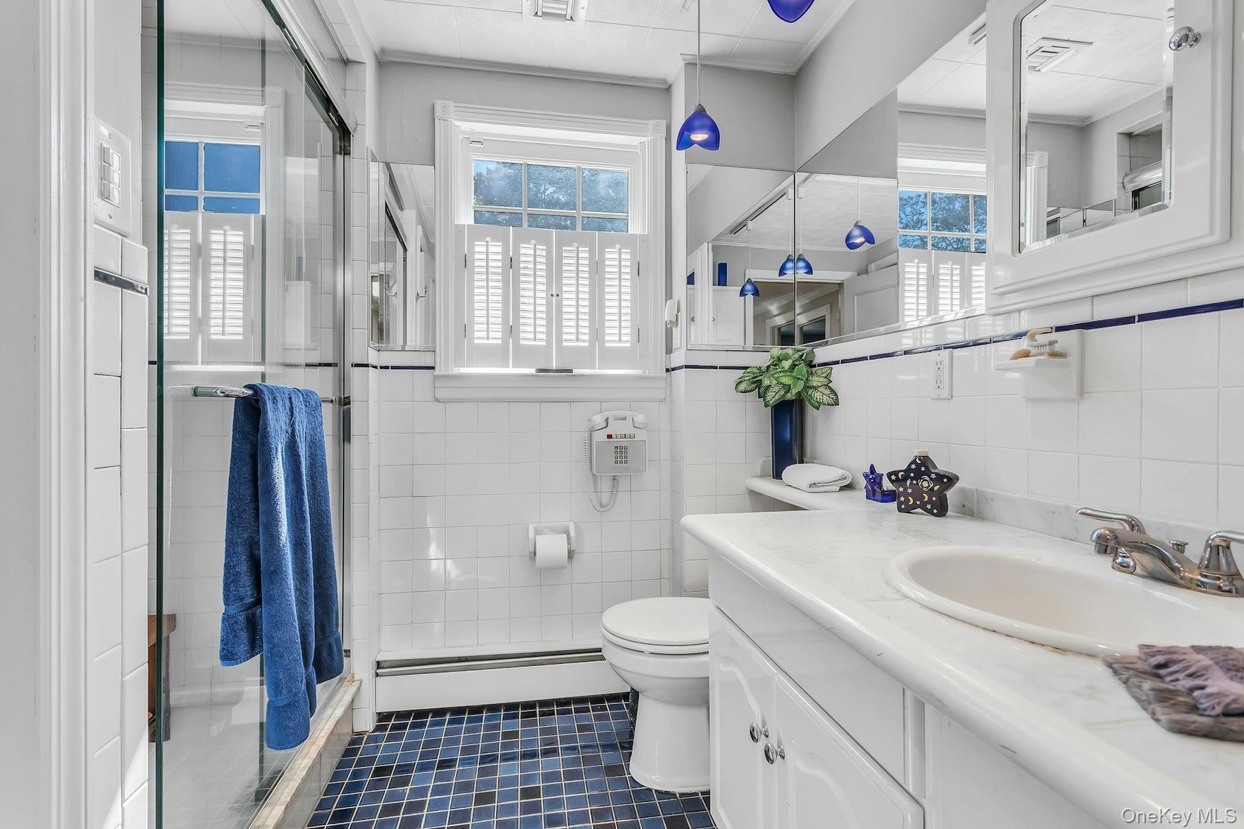 Glass-enclosed shower, dual windows with plantation shutters, white tiled walls with blue trim, built-in vanity with integrated sink, and mosaic tile 