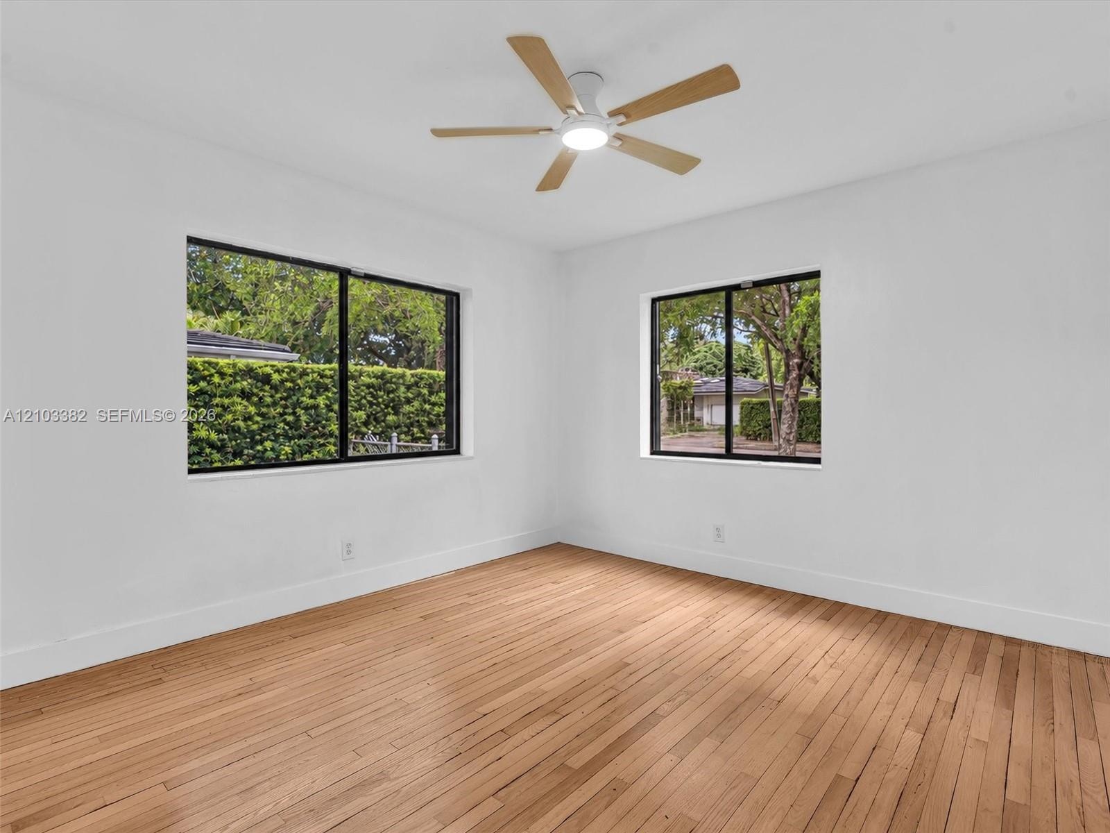 Large front/east bedroom with sunny windows. Featuring wood-finish flooring, two windows with black frames, and a multi-blade ceiling fan with integra