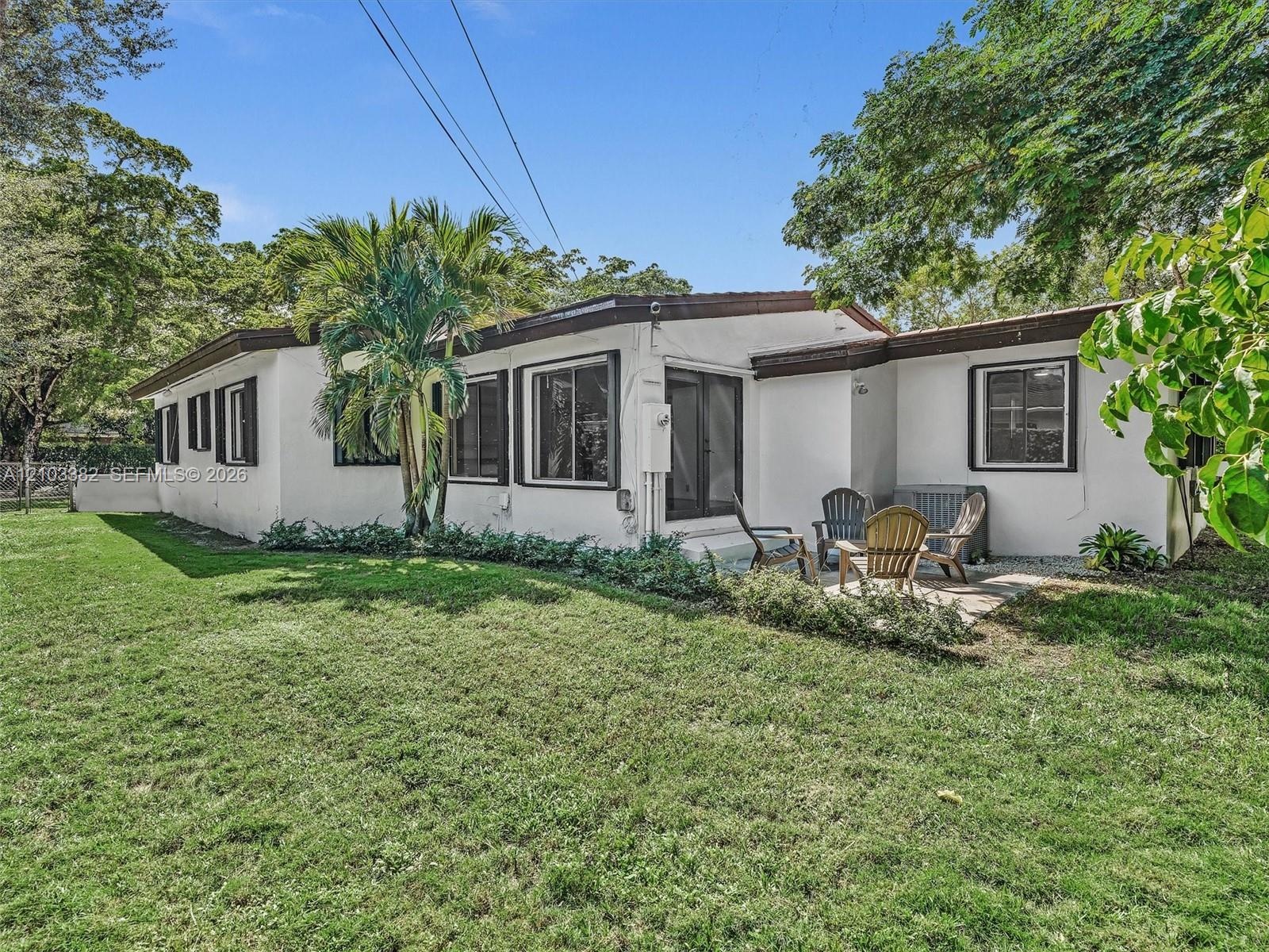 Exterior featuring white stucco siding, dark-trimmed windows, a concrete patio, and a fenced grass lawn with mature trees and palm foliage. House for 