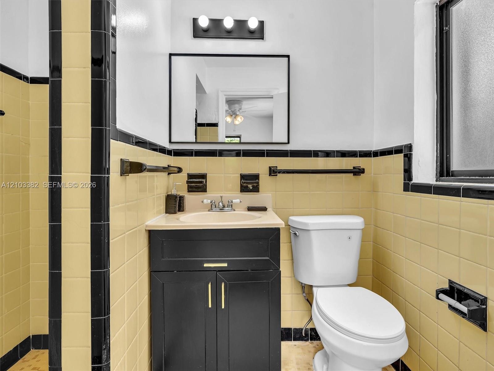Black vanity cabinet with brass-tone hardware, rectangular wall-mounted mirror, three-light fixture, and ceramic tile wall accents featuring contrasti