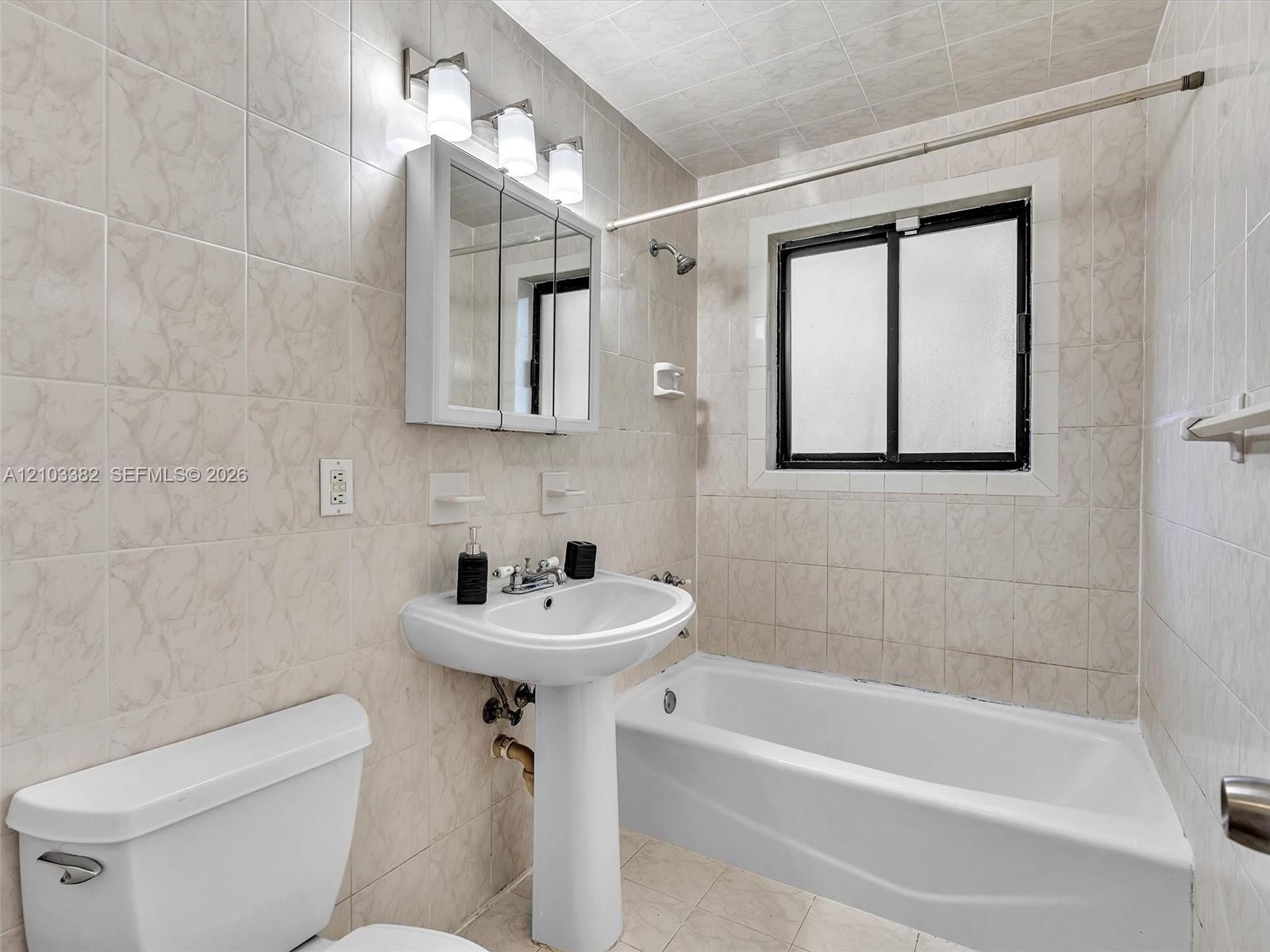 Full bathroom featuring a pedestal sink, a bathtub with a tiled surround, a mirrored medicine cabinet with integrated lighting, and a window