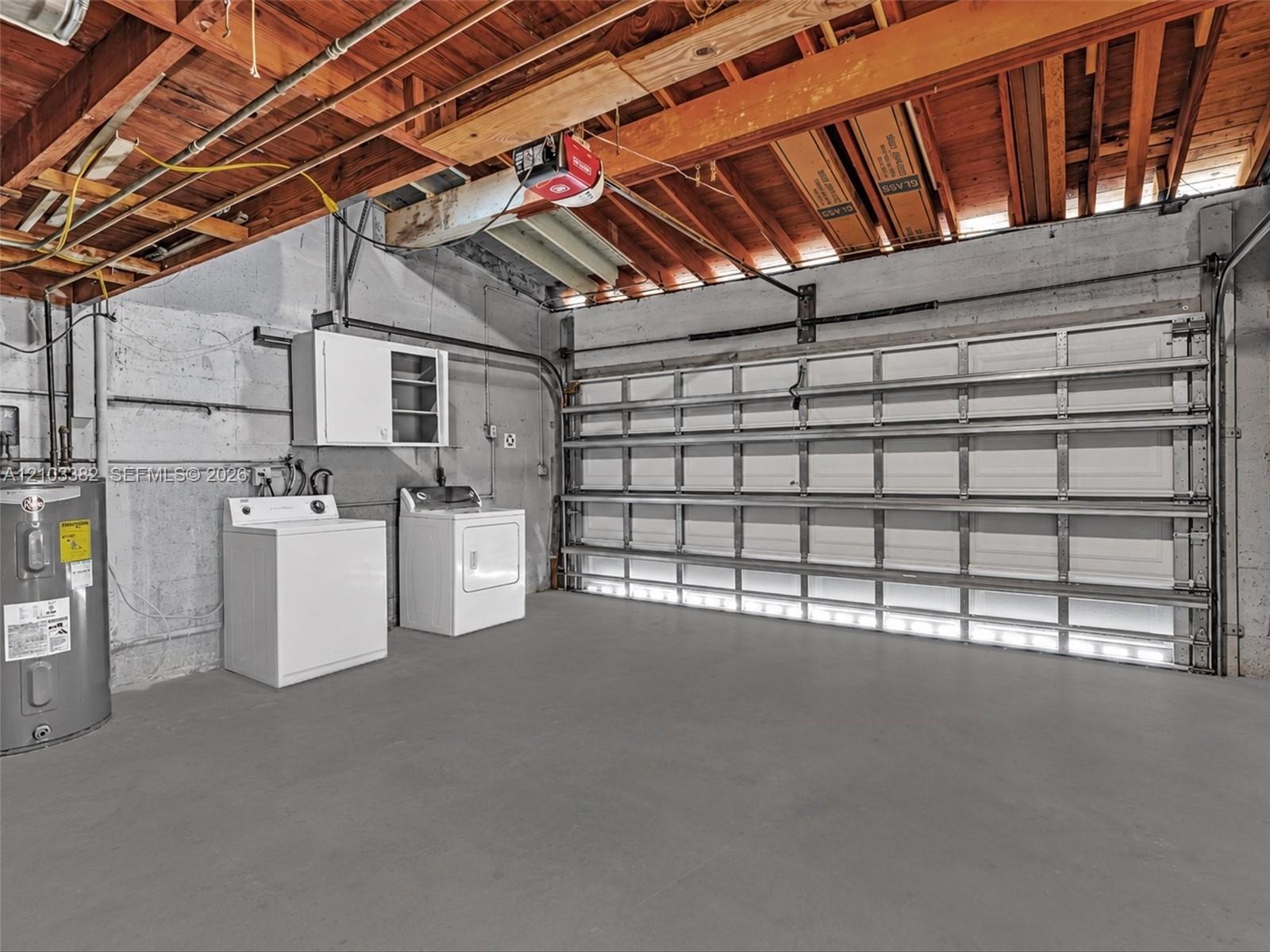 Huge 2 car, attached garage featuring a white sectional roll-up door, exposed wood joist ceiling, concrete walls, and concrete flooring. House for ren