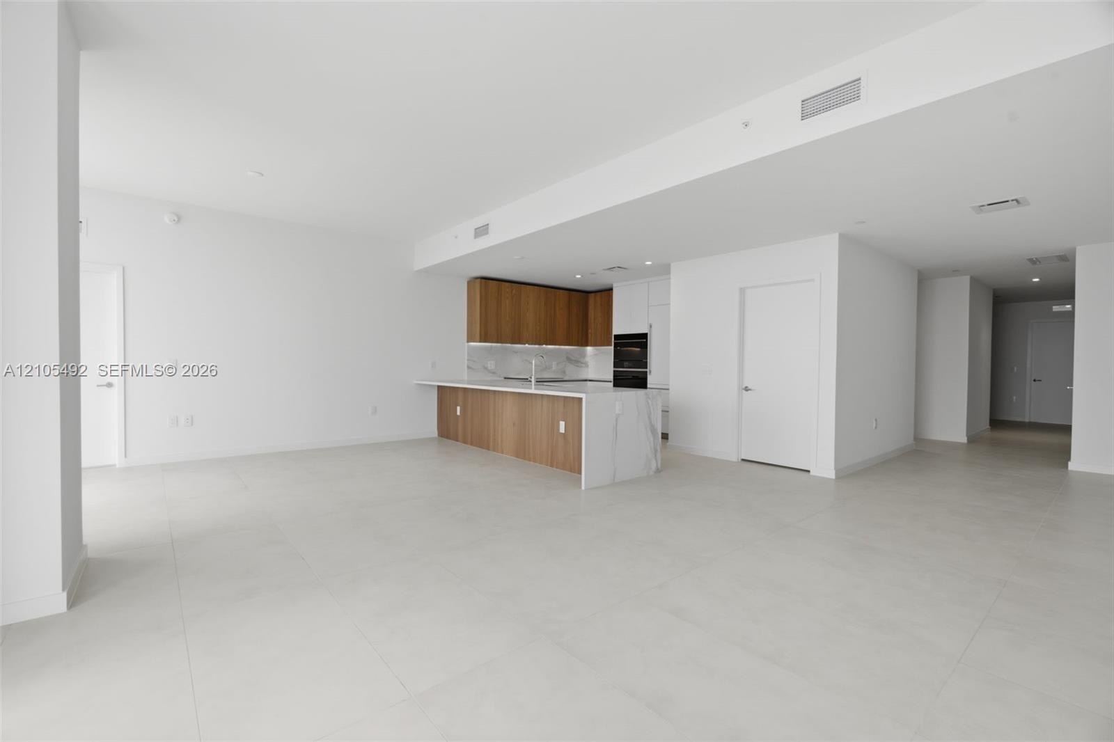 Open-concept interior featuring large-format tile flooring, wood-finish cabinetry, a waterfall-edge kitchen island, and built-in wall ovens