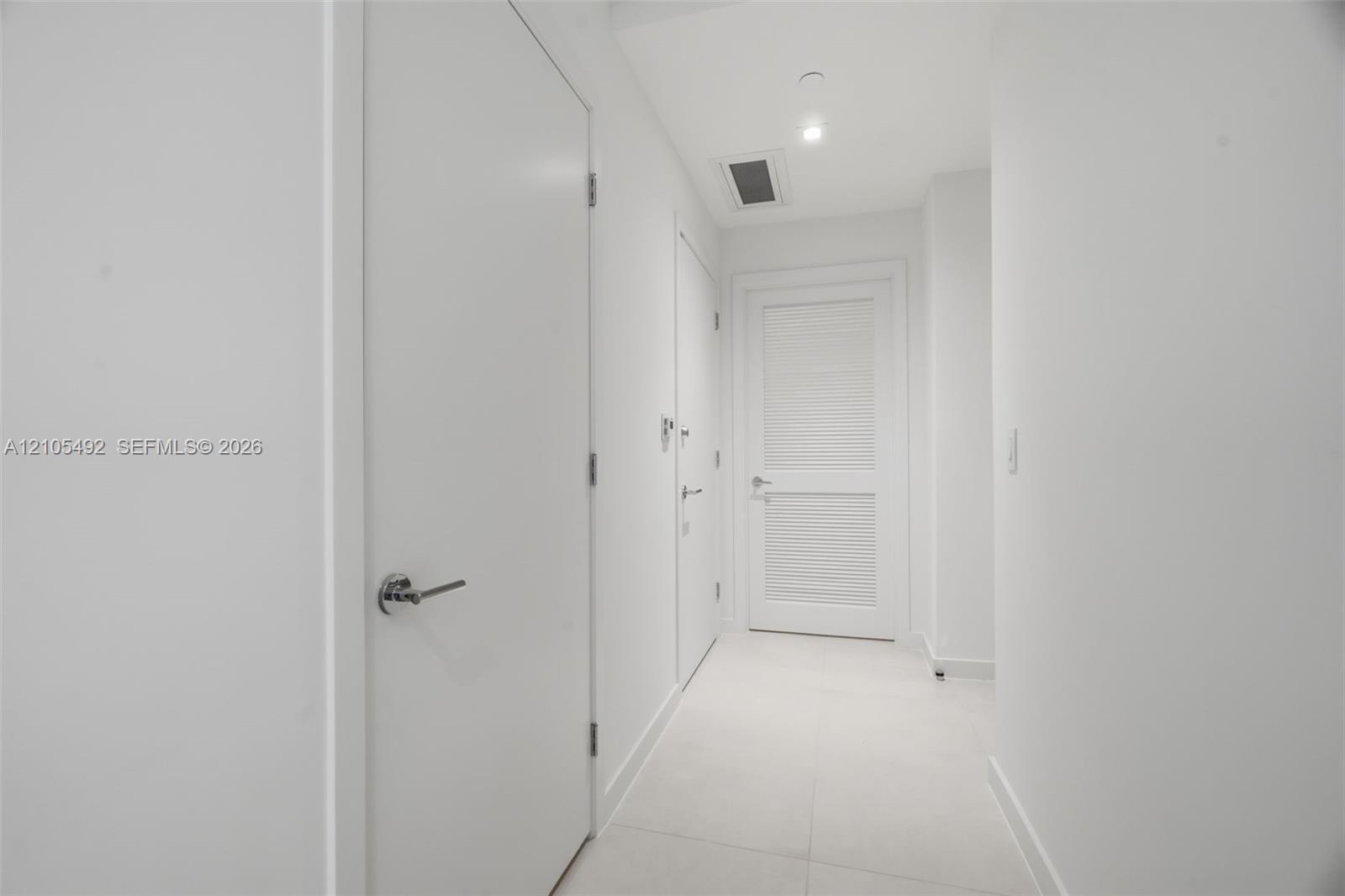 Interior corridor featuring smooth white walls, large-format tile flooring, recessed ceiling lighting, and solid-core doors with brushed-metal lever h
