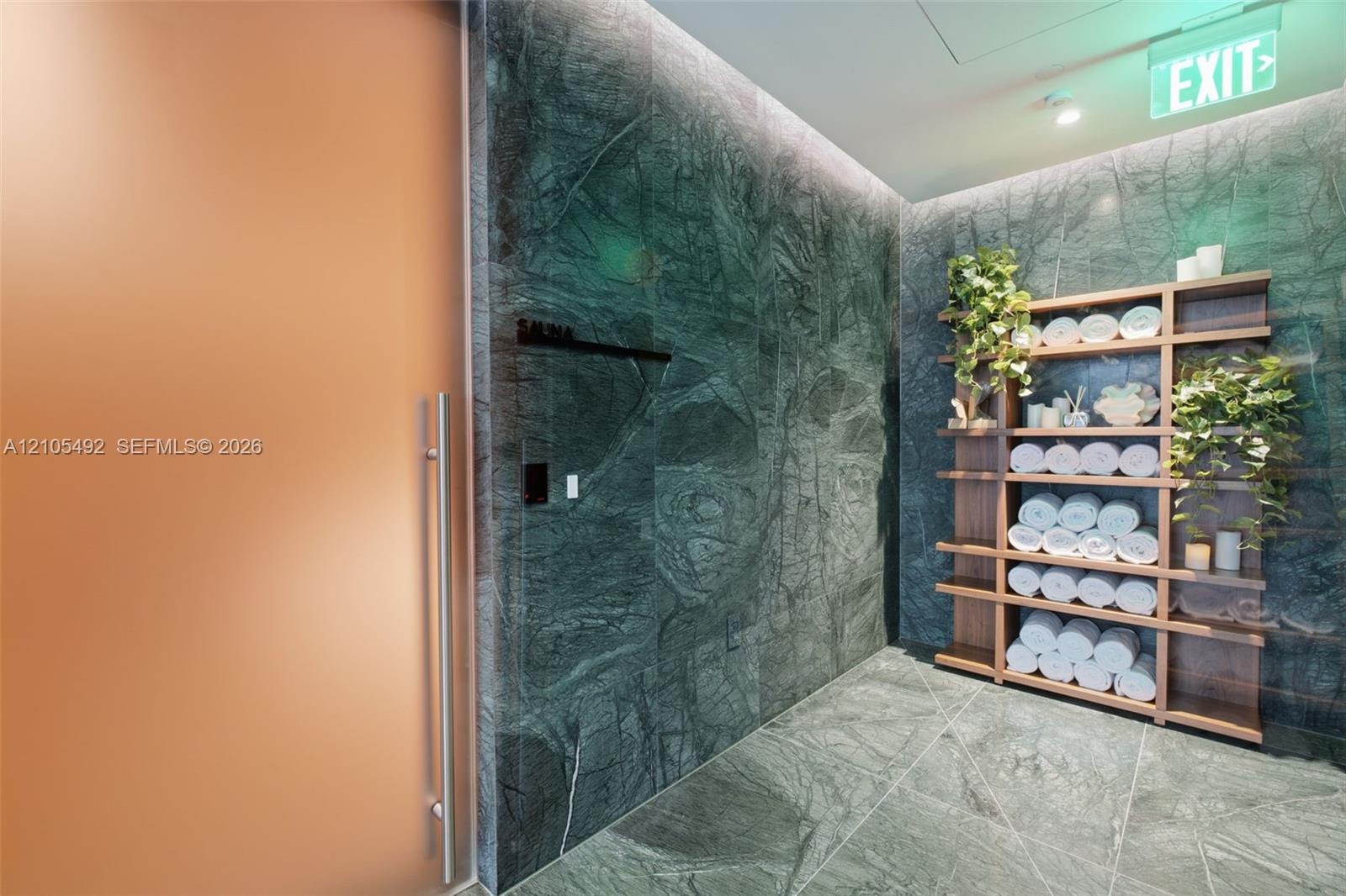 Floor-to-ceiling dark green stone wall tiling, integrated wood-finish shelving unit, oversized frosted glass door with metal pull handle, and stone ti