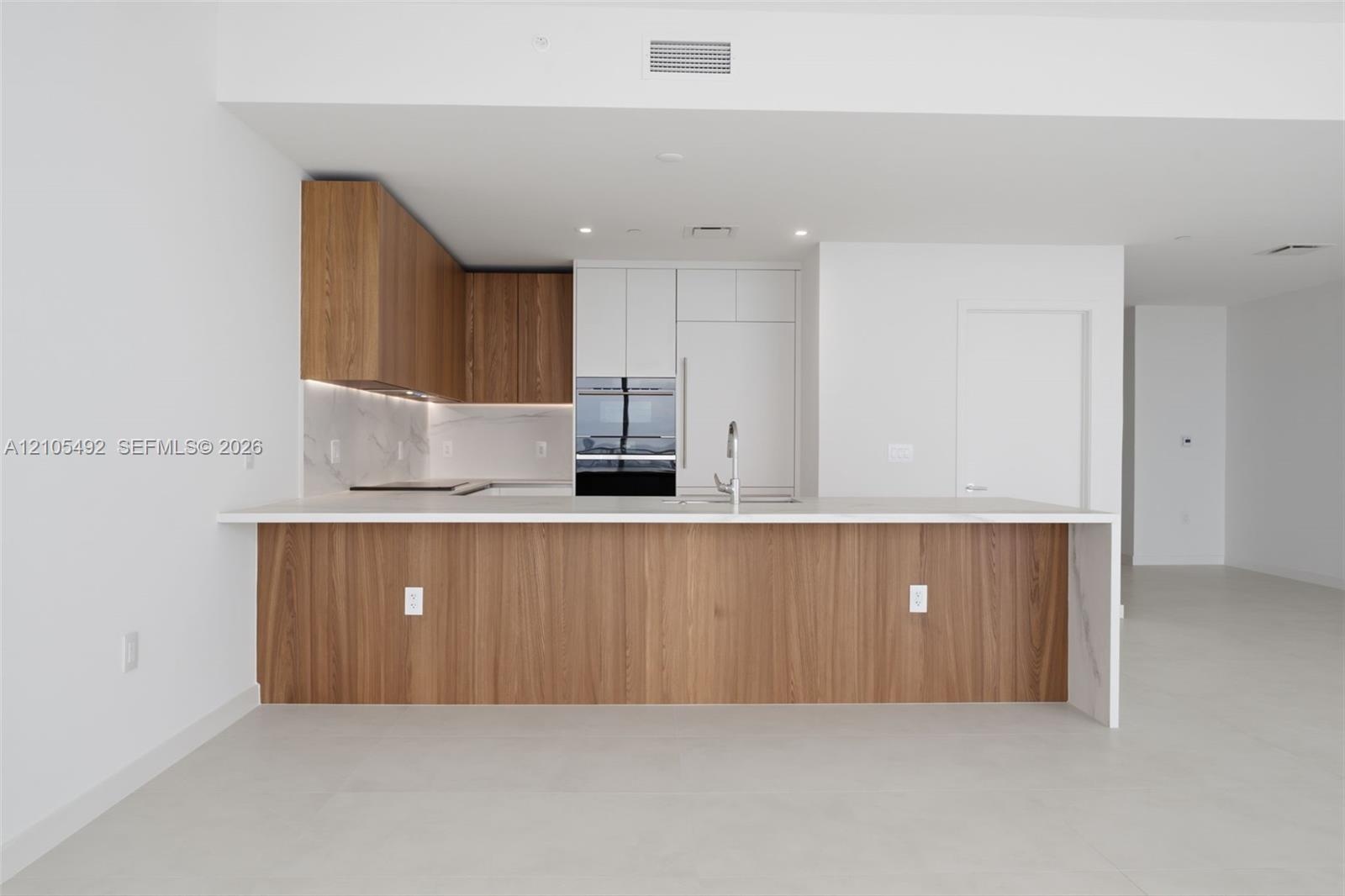 Kitchen featuring wood-finish cabinetry, white flat-panel upper cabinets, a waterfall-edge countertop, stone-finish backsplash, and built-in stainless