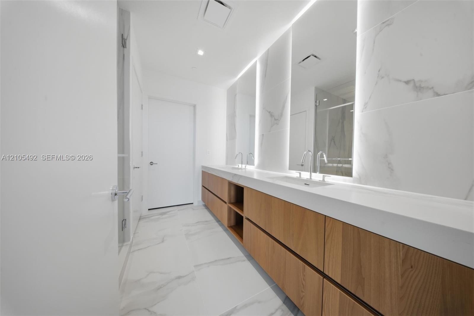Dual-sink floating vanity with wood-finish cabinetry, solid surface countertop, large-format tile wall surrounds, and integrated LED vanity mirrors