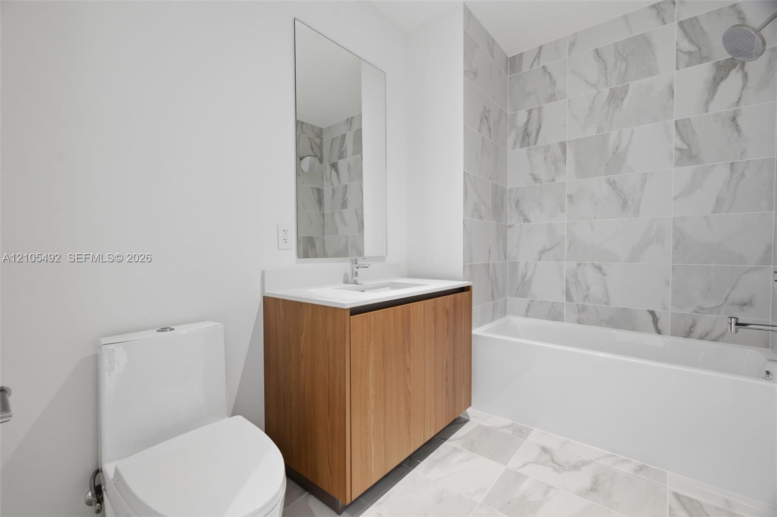 Bathroom featuring a wood-finish vanity with a white countertop, rectangular wall mirror, full-height stone-patterned tile shower surround, and white 