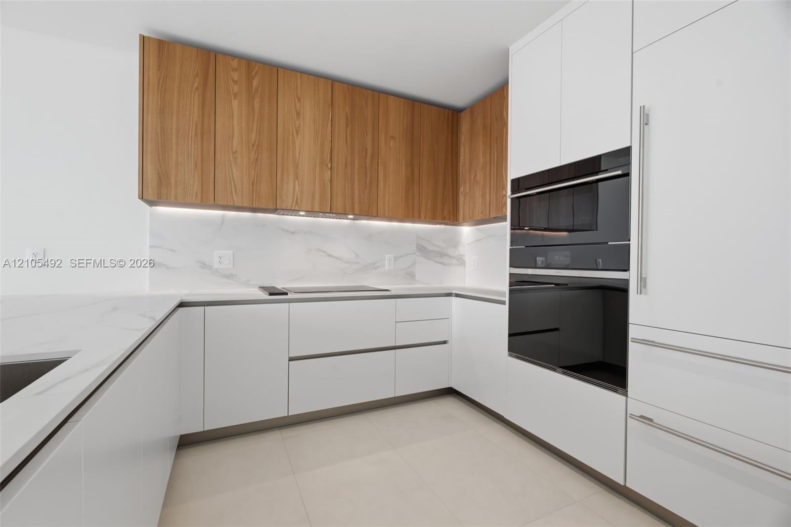 Kitchen featuring two-tone cabinetry with wood-finish upper units and white lower cabinetry, light-toned stone-finish countertops, full-height stone-f