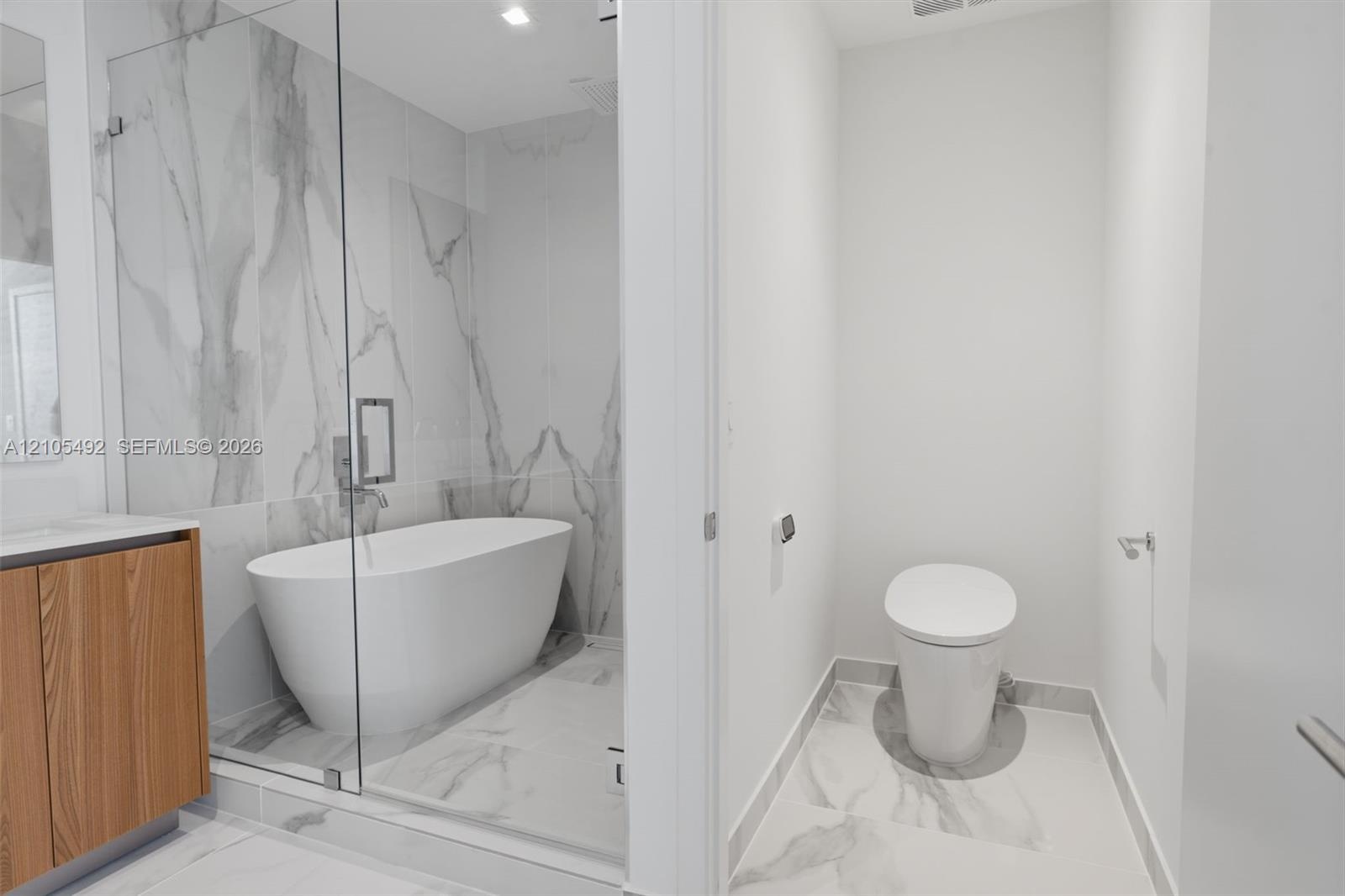 Bathroom featuring a glass-enclosed wet room with a freestanding soaking tub, wood-finish vanity cabinetry, and large-format stone-patterned tile floo
