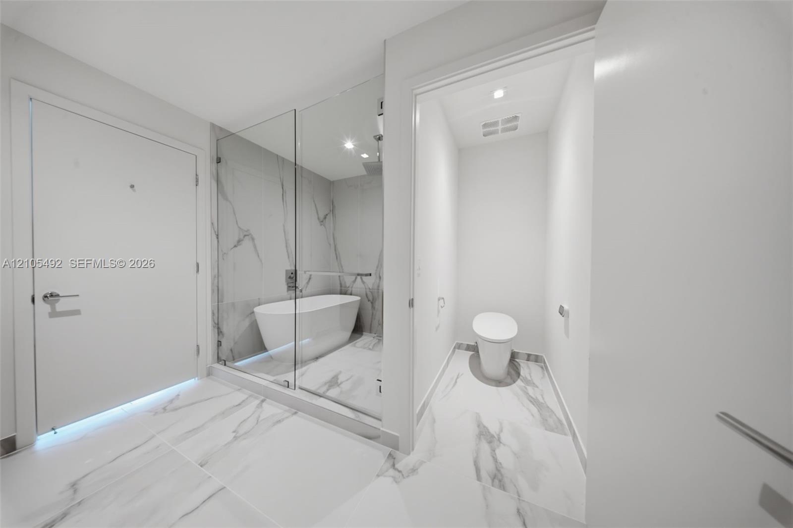 Enclosed wet room featuring a freestanding soaking tub, floor-to-ceiling glass paneling, stone-patterned tile flooring, wall-mounted shower hardware, 