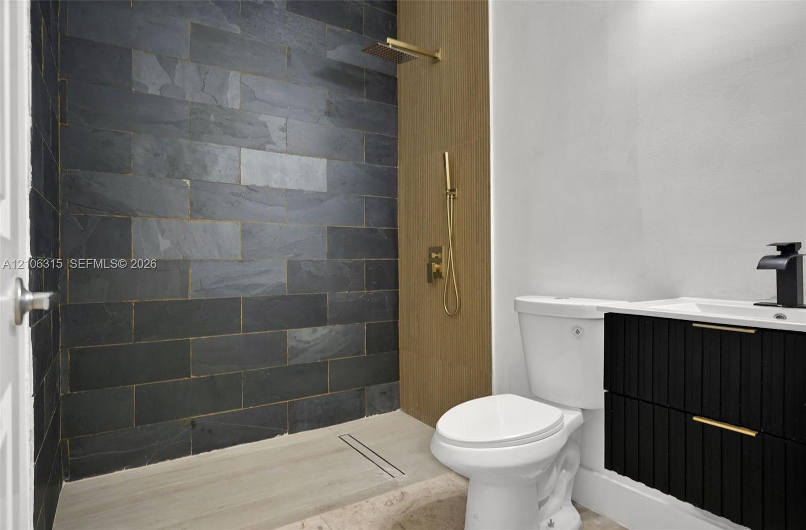Walk-in shower featuring dark stone-style tile, gold-finish hardware, and a wood-finish panel accent, paired with a black floating vanity and integrat