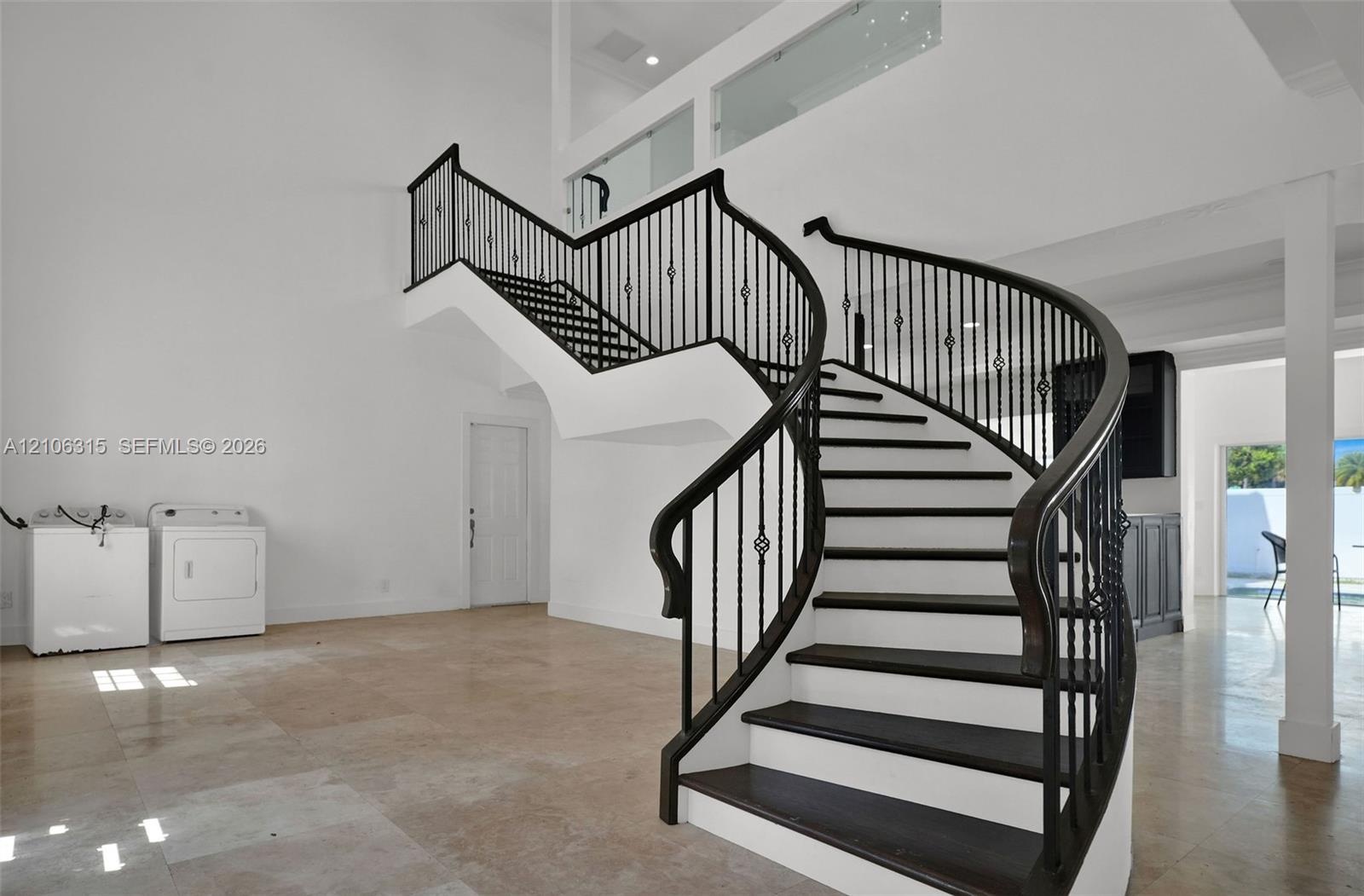 Curved staircase featuring dark wood treads and a black metal railing, complemented by double-height ceilings, stone tile flooring, and a glass-panele