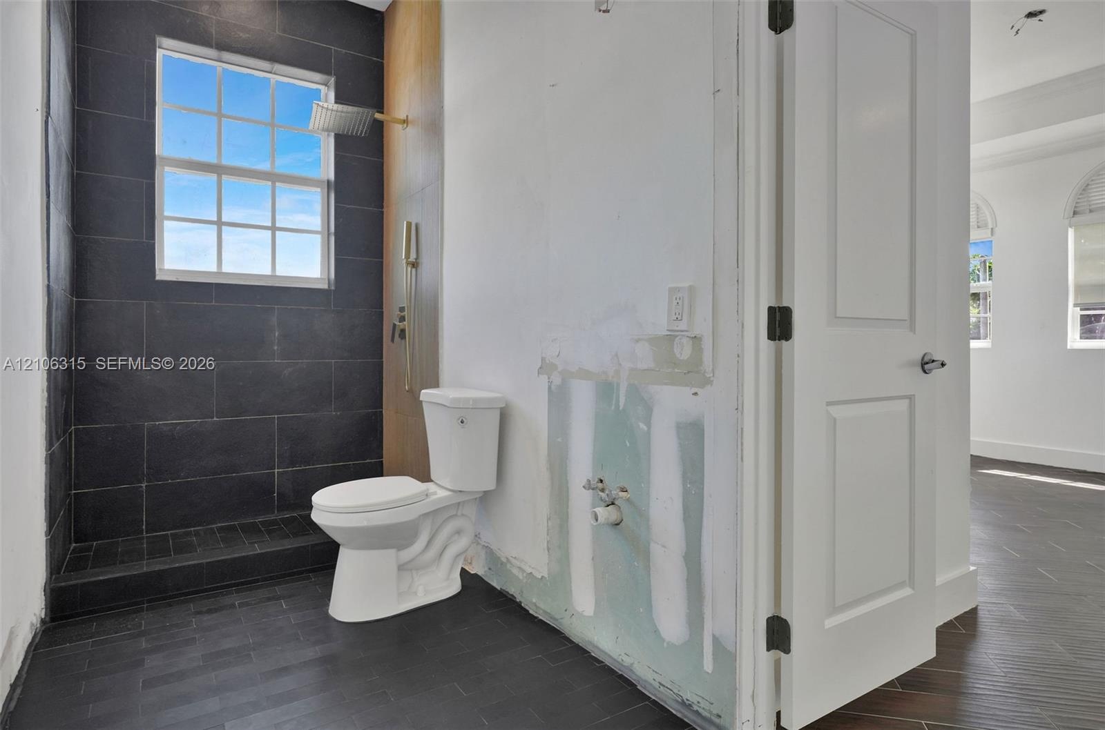 Walk-in shower enclosure with large-format dark tile, wall-mounted rain shower head, dual-pane grid window, and wood-finish flooring