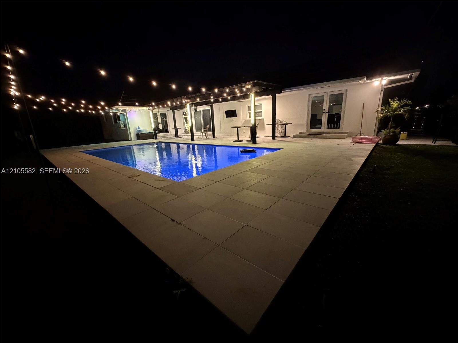 In-ground pool surrounded by a large paver patio, featuring string lighting and a covered pergola