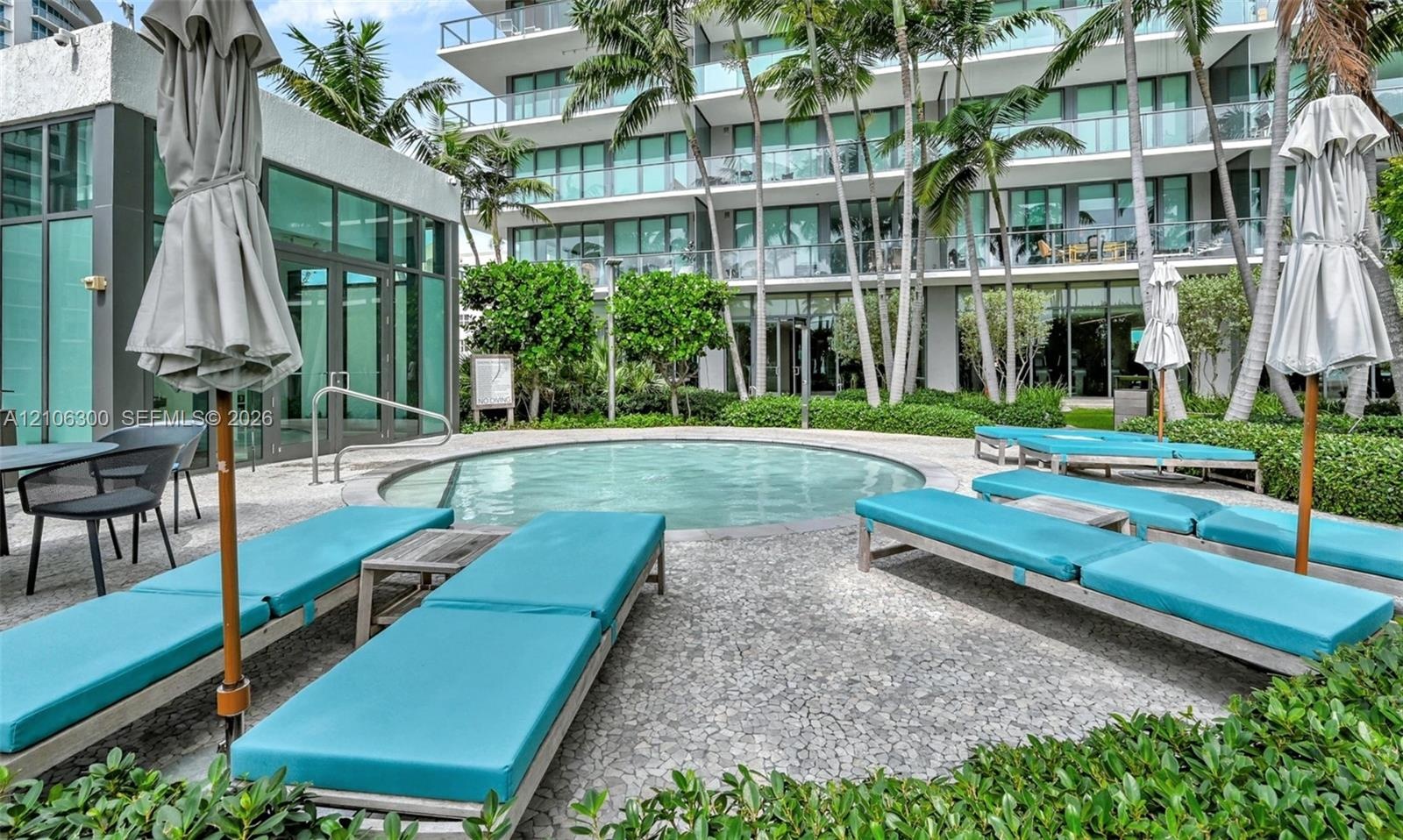 Circular swimming pool surrounded by a stone-paved deck, landscaped garden beds, and palm trees