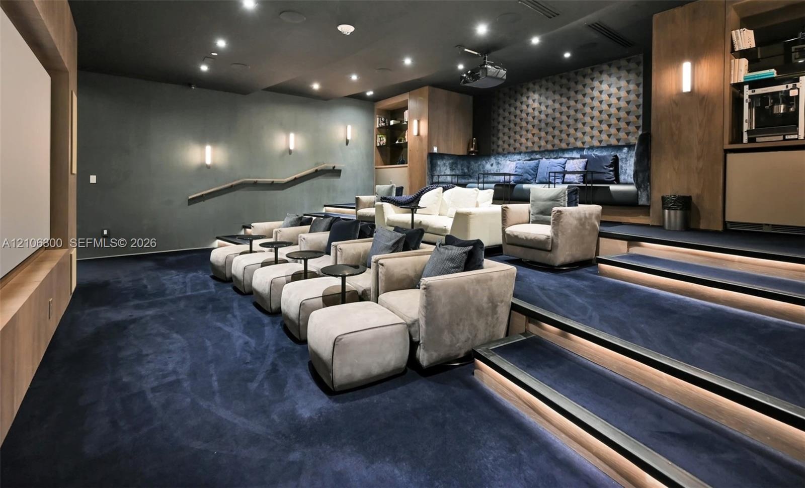 Media room featuring tiered flooring, recessed stair lighting, integrated wooden cabinetry, a projector, and wall-mounted sconce lighting