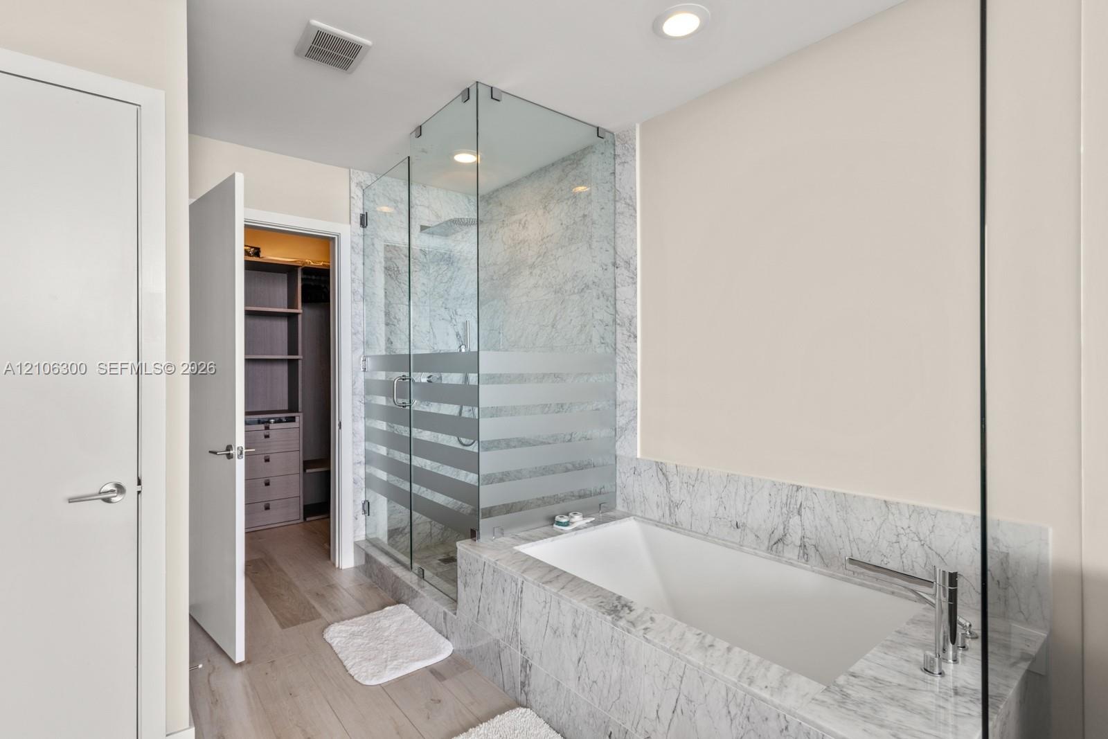 Stone-surround soaking tub with deck-mounted faucet, glass-enclosed shower, wood-finish flooring, and walk-in closet with built-in cabinetry