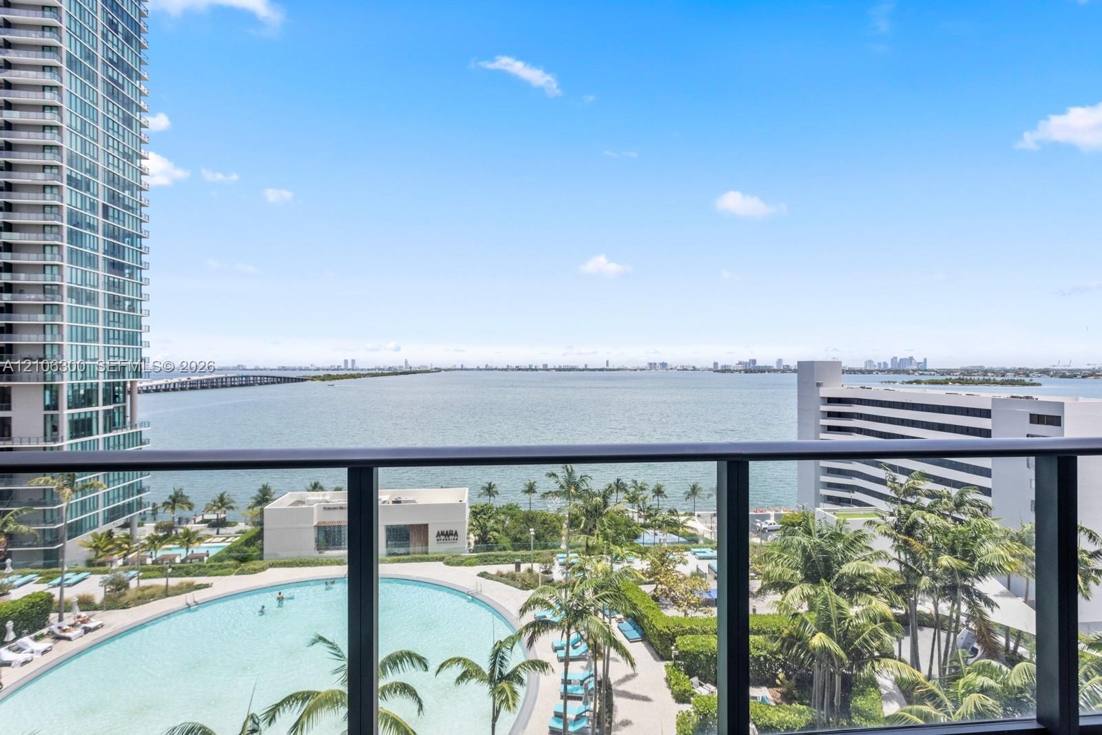 Balcony featuring a dark metal railing, overlooking a large swimming pool, landscaped grounds with mature palm trees, and coastal water