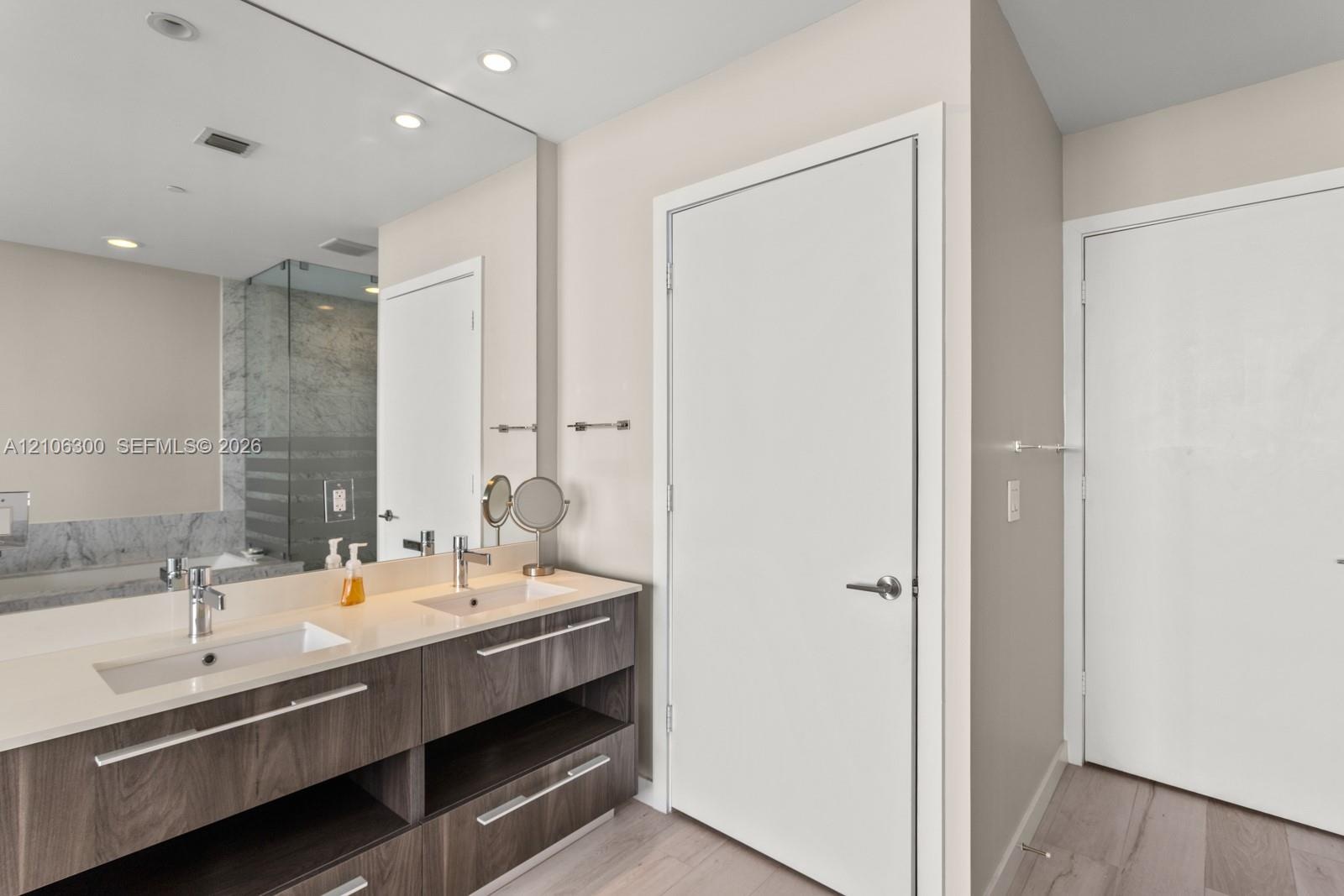 Double vanity featuring a light-toned countertop with integrated dual sinks, wood-finish cabinetry with linear hardware, full-width wall mirror, and r