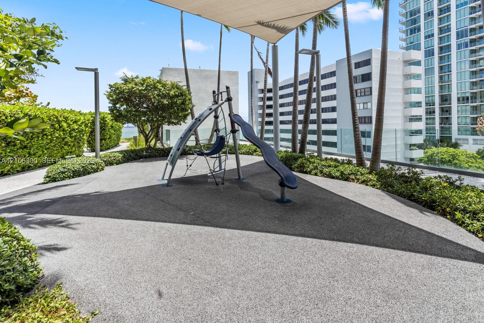 Outdoor recreation area featuring a play structure with a slide, rubberized surfacing, overhead shade canopy, perimeter landscaping, and vertical ligh