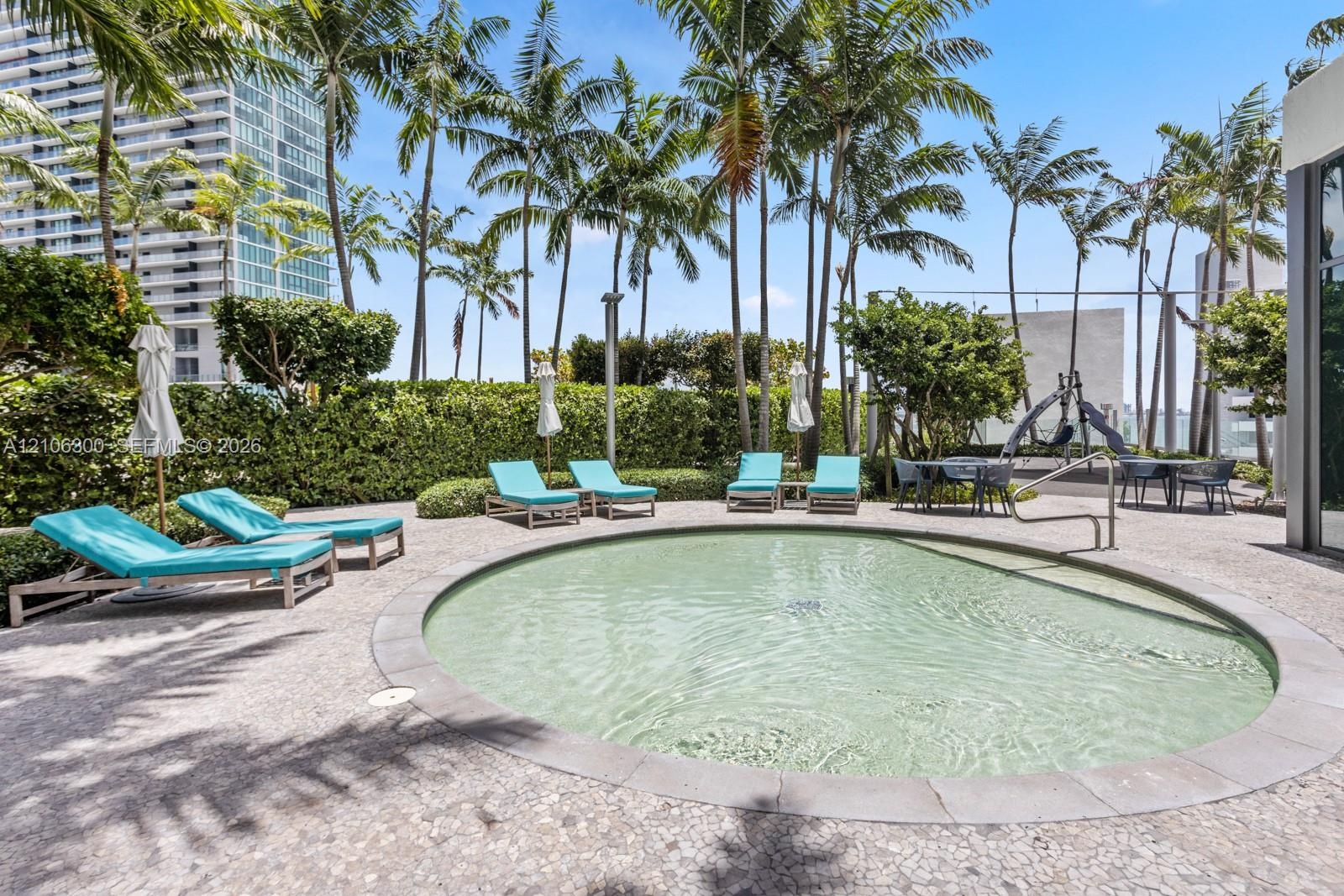 Circular swimming pool featuring a stone-tile border, set within a paved deck with mature palm trees and manicured privacy hedging
