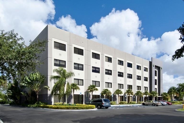 Four-story multi-unit structure featuring a concrete facade, grid-patterned exterior, glass window panels, and integrated asphalt surface parking