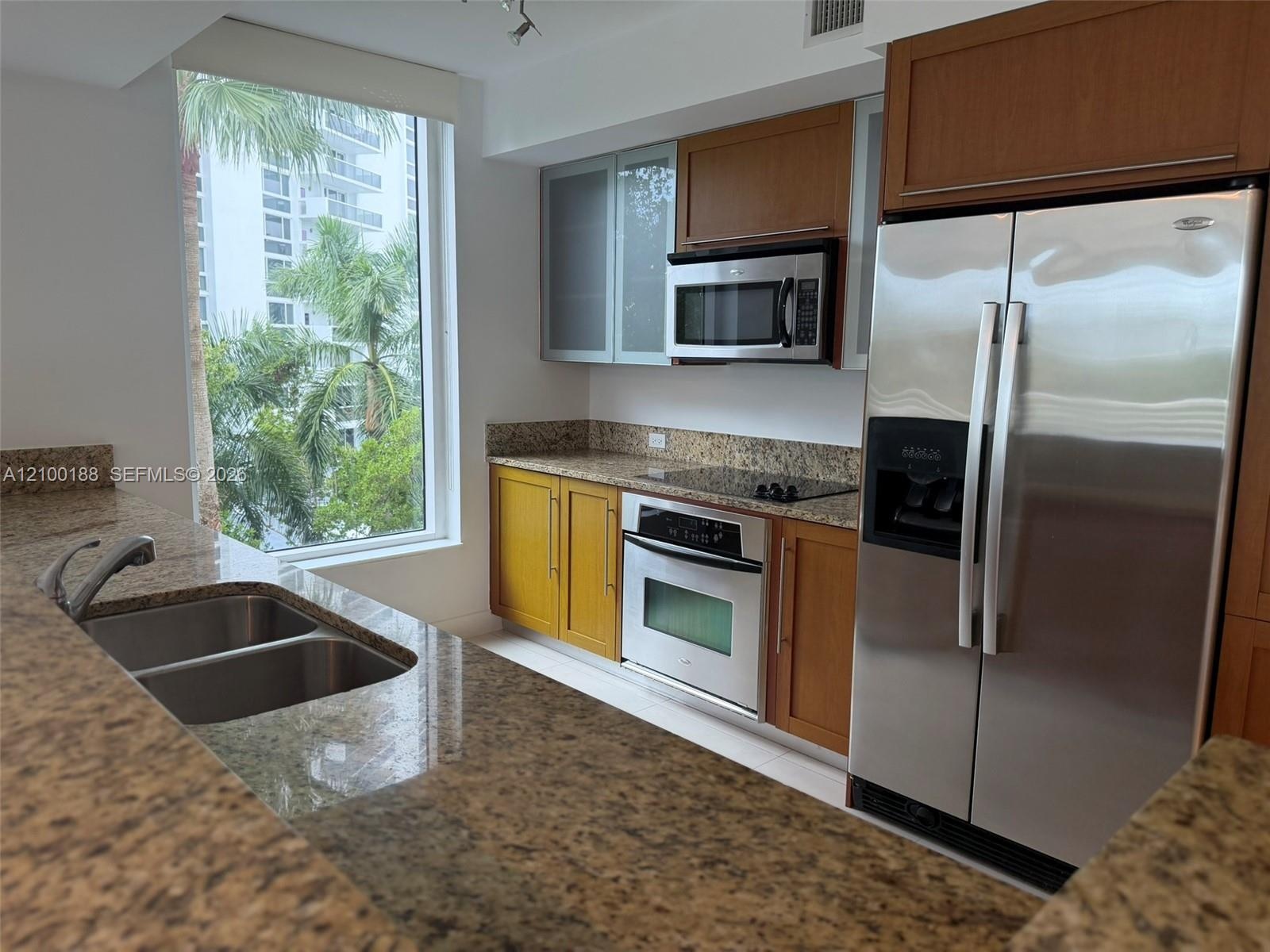 Kitchen featuring stone countertops, stainless steel appliances, wood cabinetry, dual-basin sink with pull-down faucet, and floor-to-ceiling window