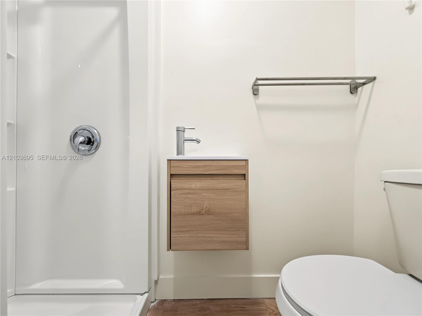 Floating wood-finish vanity with a white countertop and single-handle faucet, integrated shower stall with chrome fixtures, and a wall-mounted metal t