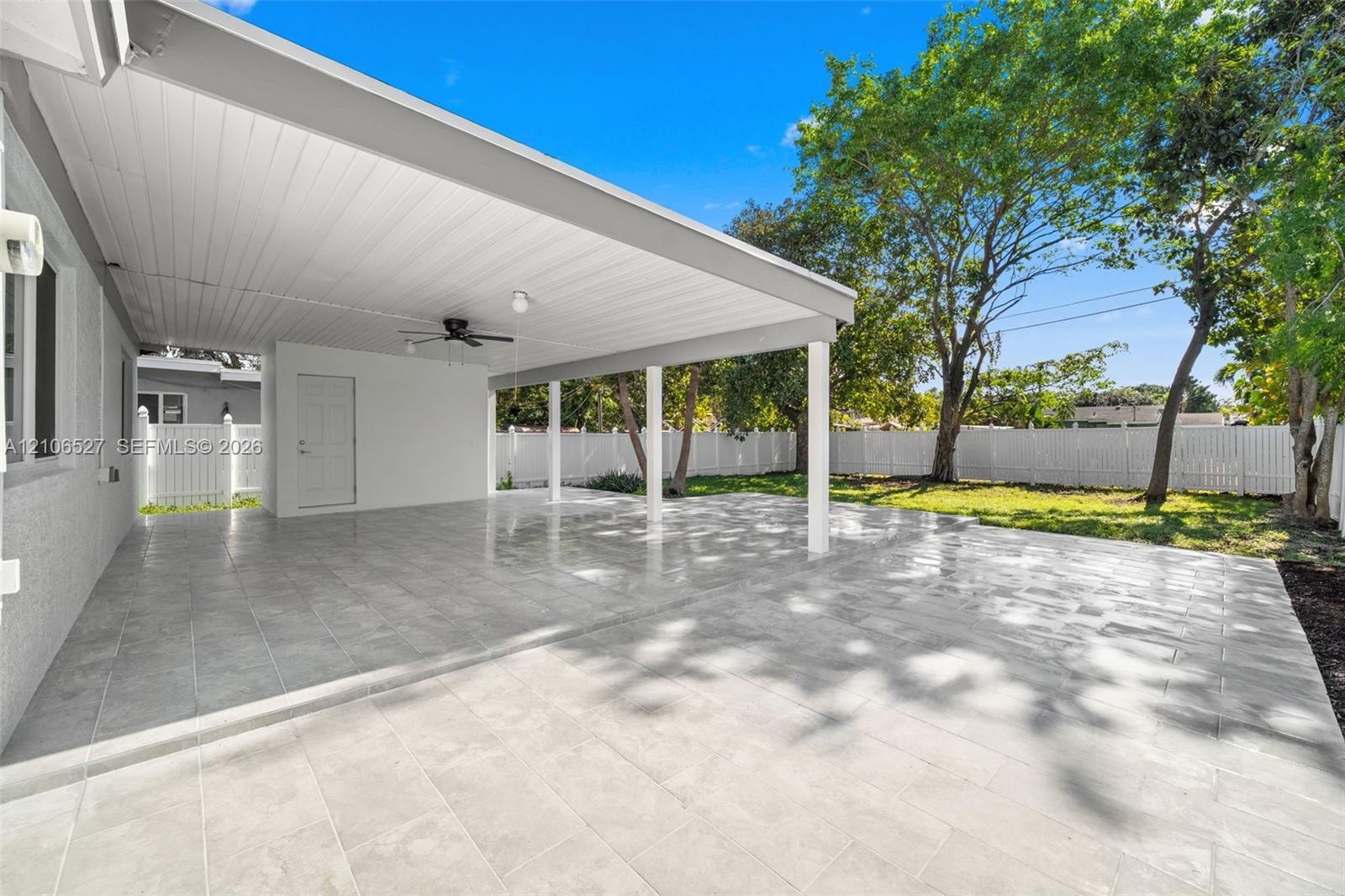 Covered patio featuring ceramic tile flooring, a ceiling fan, white vinyl perimeter fencing, and mature landscaping