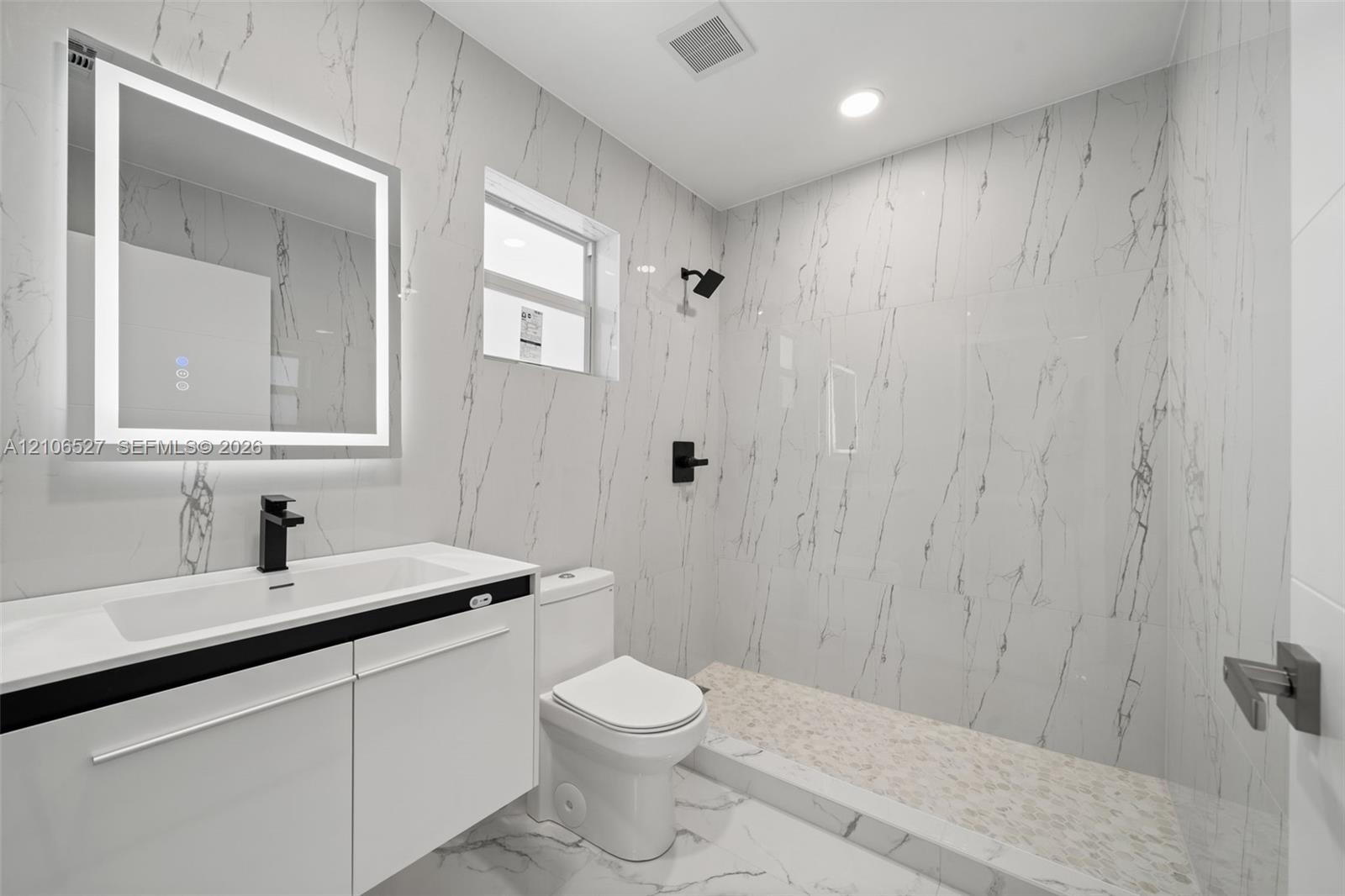 Bathroom featuring full-height stone-finish wall tile, walk-in shower with pebble-stone flooring, integrated LED vanity mirror, white cabinetry with a