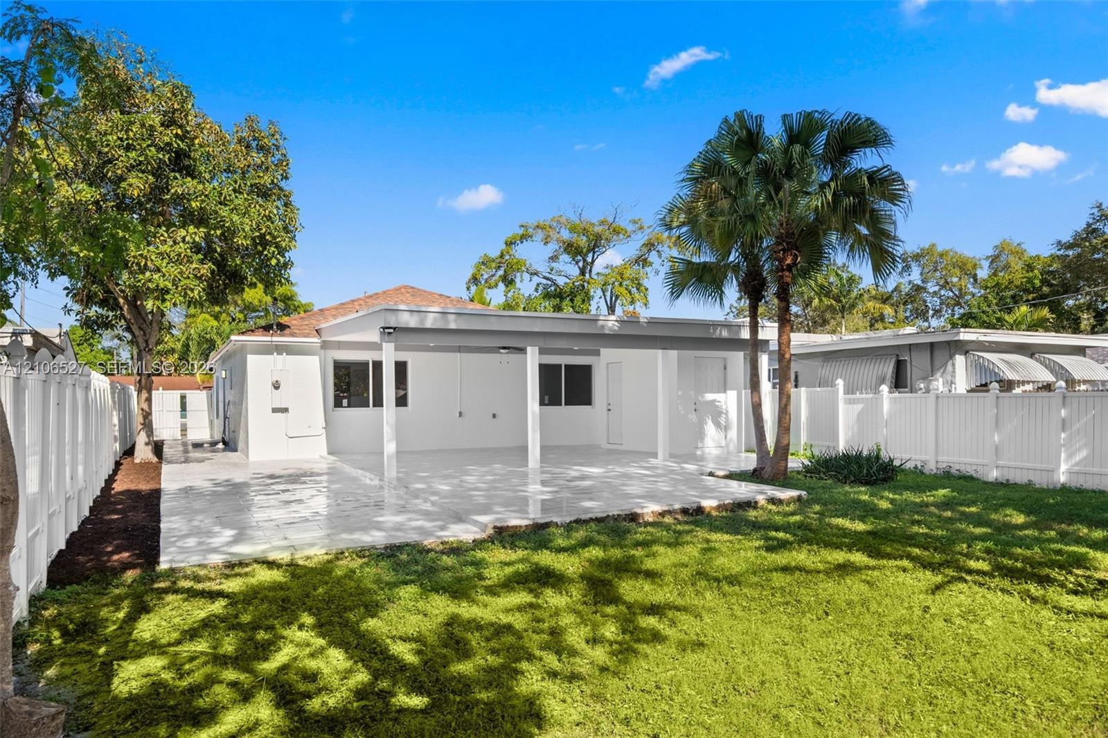 Rear exterior featuring a covered patio, paved tile flooring, enclosed white vinyl fencing, and palm trees