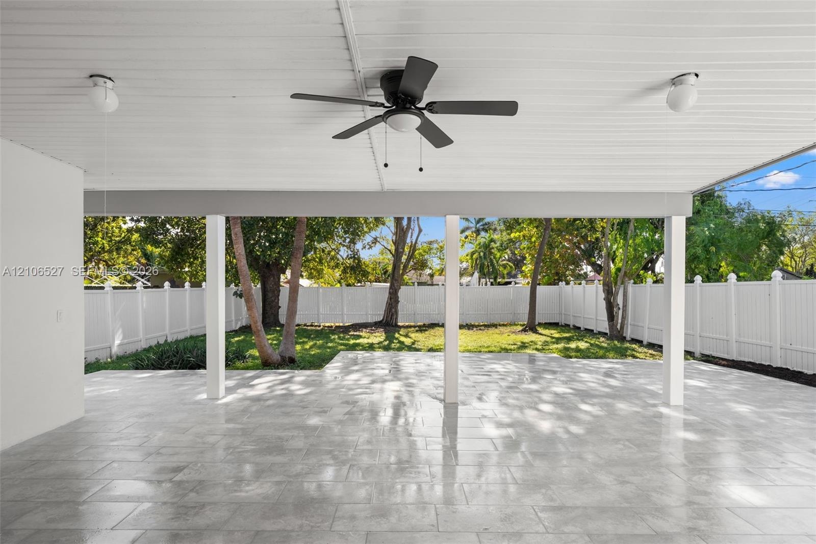 Covered patio featuring stone-finish flooring, a ceiling fan, dual overhead light fixtures, and a white vinyl privacy fence surrounding the rear groun