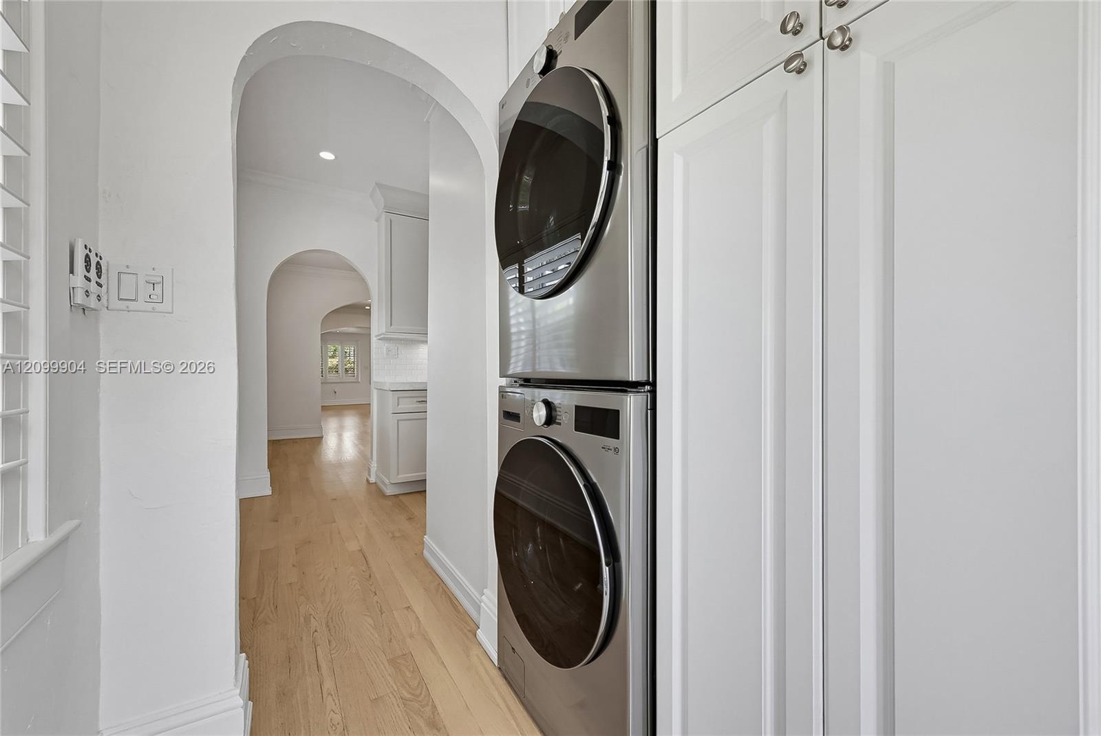 Stacked stainless steel laundry appliances, floor-to-ceiling cabinetry, wood-finish flooring, and arched architectural openings