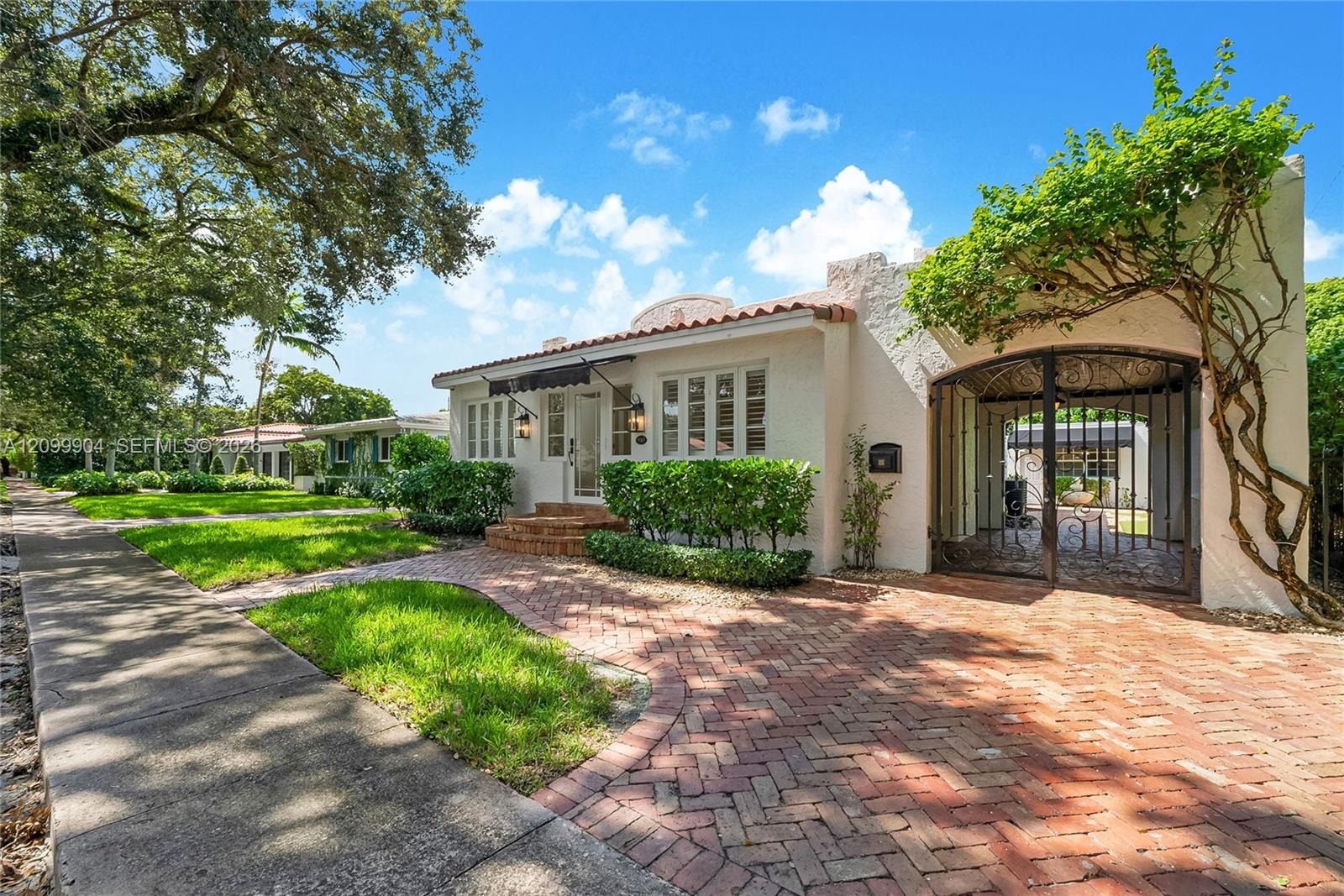 Stucco exterior featuring mission-style architecture, a red brick herringbone driveway, terracotta roof accents, and a decorative wrought iron gate