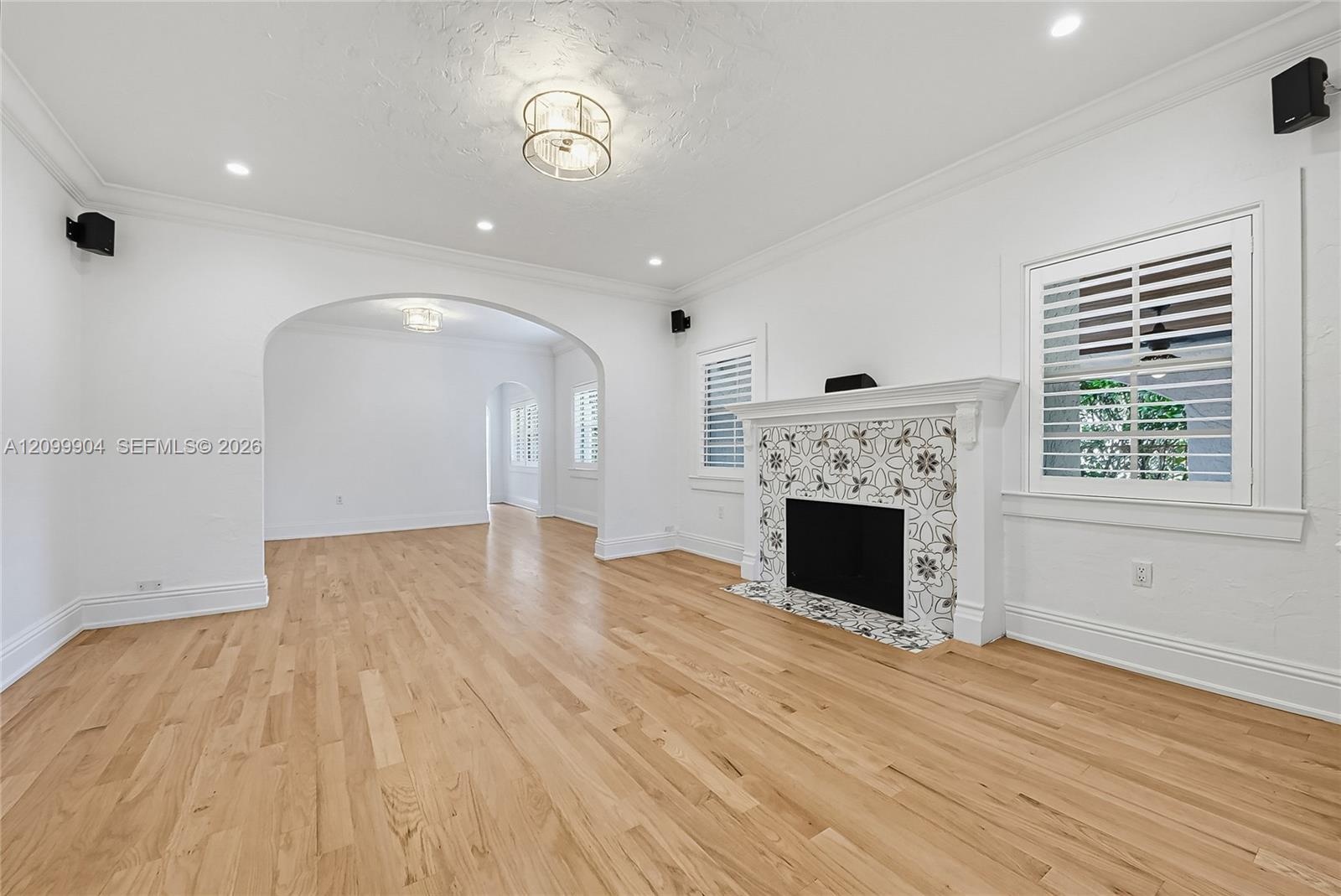 Living area featuring wood-finish flooring, a decorative tile-surround fireplace, arched architectural entryways, crown molding, and plantation shutte