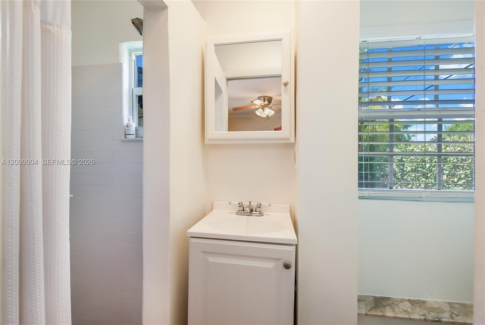 White vanity with an integrated basin, chrome-finished dual-handle faucet, white framed medicine cabinet, and a shower area featuring white wall tile